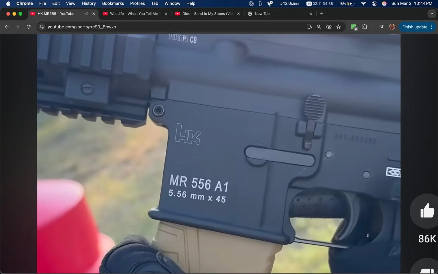 The image displays a close-up view of a firearm, specifically the lower receiver of a rifle. The firearm is marked with the logo "HK," which stands for Heckler & Koch, a well-known firearms manufacturer. The model designation "MR 556 A1" is prominently displayed in white text on the side of the receiver. Below this, the caliber is indicated as "5.56 mm x 45," also in white text. 

The lower receiver features a textured grip area, which is likely designed for better handling. There is a safety selector switch visible, indicating the firearm's operational mode, and a magazine release button is also present. The overall color of the firearm appears to be a matte black, which is common for tactical firearms.

The background is slightly blurred, but hints of green grass and a red object, possibly a container or a piece of equipment, can be seen. The image suggests an outdoor setting, likely a shooting range or training area, given the context of the firearm being displayed.

The hand holding the firearm is gloved, indicating a focus on safety and grip control. The glove appears to be tactical in nature, possibly made of a durable material suitable for handling firearms. The focus of the image is on the firearm itself, with no visible people or additional objects in the immediate foreground. 

Overall, the image captures a detailed view of the firearm's features and markings, set against a natural outdoor backdrop.