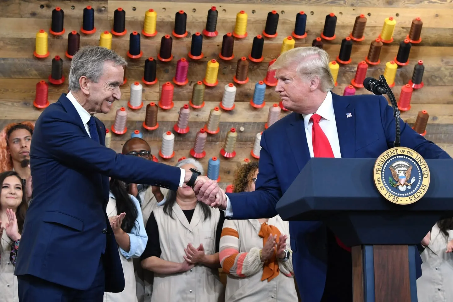 The image depicts two men engaged in a handshake at a formal event. The man on the left is dressed in a dark suit with a light blue shirt and a tie, appearing to smile as he shakes hands. He has short, light-colored hair and is looking at the man on the right. The man on the right is wearing a dark blue suit with a white shirt and a red tie. He has light-colored hair styled back and is looking slightly to the side, with a serious expression on his face.

In the background, there is a wooden wall adorned with colorful spools of thread, arranged in a pattern. The spools are primarily in shades of red, yellow, brown, and blue, creating a visually striking backdrop. 

Behind the two men, there are several individuals, some of whom are clapping. They are dressed in beige or light-colored clothing, suggesting a uniform or coordinated attire. The expressions of the people in the background range from smiles to applause, indicating a supportive atmosphere.

A podium is visible in the foreground, featuring the presidential seal of the United States, indicating that this is a formal event likely involving a public figure. The setting appears to be indoors, possibly in a workshop or studio environment, given the presence of the spools of thread and the casual attire of the audience members. 

Overall, the scene conveys a moment of interaction between two individuals, set against a backdrop that emphasizes creativity and craftsmanship.