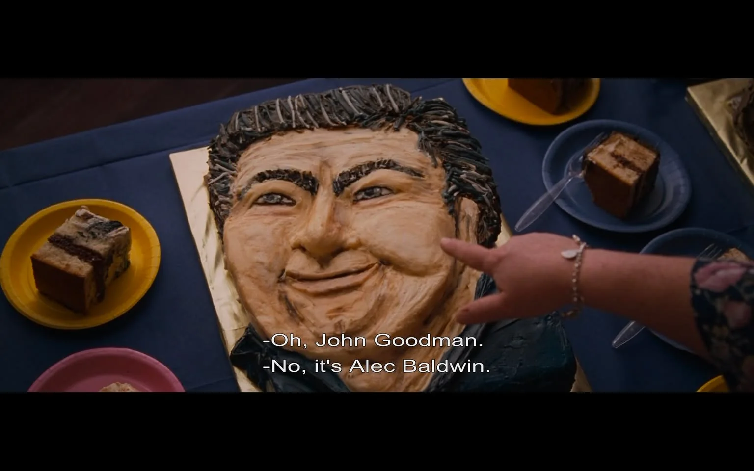 The image depicts a cake designed to resemble a person's face, prominently displayed on a blue tablecloth. The cake features a detailed sculpted face with a smiling expression, characterized by thick, dark hair styled upwards, and pronounced facial features including a broad nose and full cheeks. The cake is decorated with various shades of icing to create depth and texture, particularly around the hair and facial contours.

In the foreground, a hand is pointing at the cake, suggesting interaction or discussion about its likeness. The hand is adorned with a bracelet and appears to belong to a person wearing a floral-patterned sleeve, indicating a casual or festive setting.

Surrounding the cake are several plates with slices of cake, including a rectangular piece with a chocolate layer and a lighter-colored piece, both resting on yellow and blue plates, respectively. The background is blurred, but it suggests an indoor environment, possibly a gathering or celebration, given the presence of food items.

Text at the bottom of the image indicates a conversation, with one person mistakenly identifying the cake as resembling John Goodman, while another corrects them, stating it resembles Alec Baldwin instead. This dialogue adds context to the scene, indicating that the cake is intended to represent a well-known figure, although the specific identity is not confirmed.

Overall, the image captures a lighthearted moment centered around a creatively designed cake, with elements of humor and social interaction evident in the dialogue. The setting appears to be informal, likely at a party or celebration.