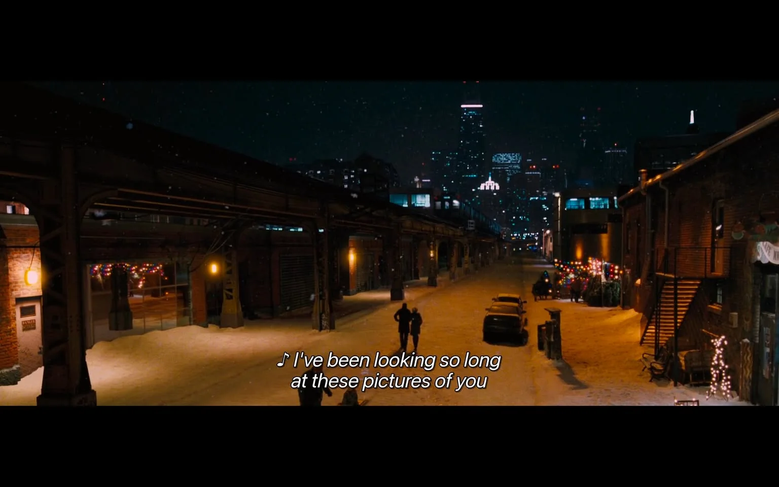 The image depicts a nighttime urban scene, likely set in a city with a skyline featuring tall buildings. The foreground shows a snow-covered street, illuminated by warm yellow lights from street lamps and nearby buildings. The ground is blanketed in snow, creating a serene winter atmosphere. 

On the left side, there is a brick building with a large window displaying colorful holiday lights, suggesting a festive ambiance. The building has a door with a small sign next to it, but the text is not readable. 

In the center of the image, two figures are walking down the street, one appearing to be in a dark coat and the other in a lighter outfit. They are positioned slightly apart, with their backs to the viewer, and seem to be engaged in conversation or simply enjoying the moment. 

To the right, there are parked cars, including a yellow vehicle and a darker one, both partially covered in snow. The street is lined with buildings that have various architectural styles, some featuring brickwork and others with modern glass facades. 

In the background, the skyline is visible with several skyscrapers, some of which have lights illuminating their outlines. Notably, one building has a distinctive spire, and another has a bright, colorful display, possibly an advertisement or decorative lighting. 

The sky is dark with hints of falling snowflakes, adding to the wintery feel of the scene. Overall, the image captures a quiet, picturesque moment in a bustling urban environment during winter.