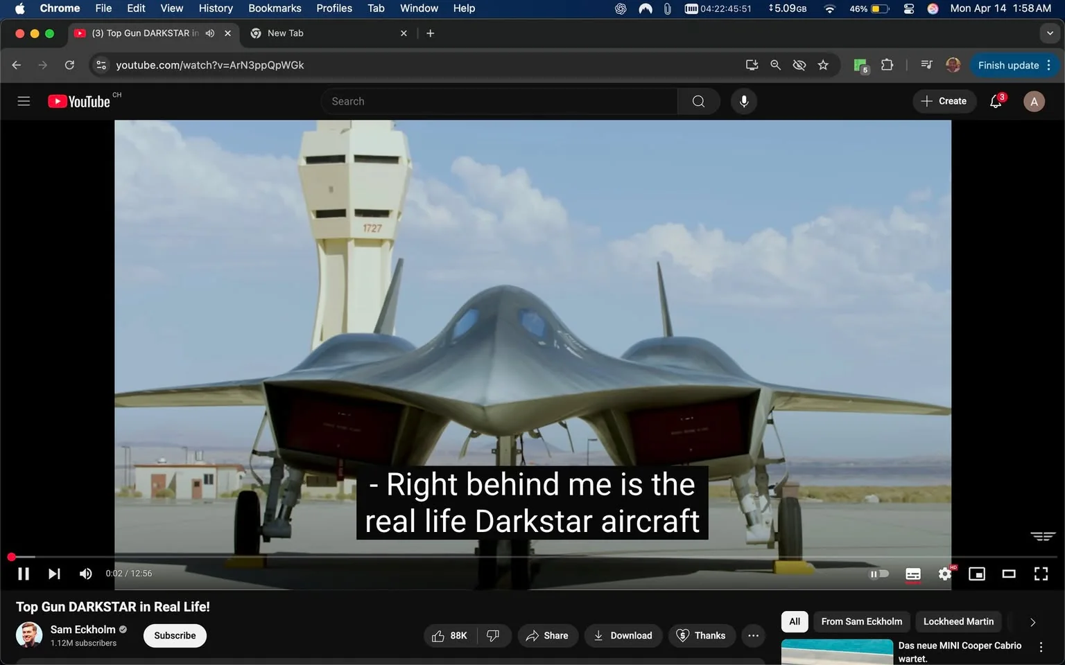 The image features a scene from a YouTube video titled "Top Gun DARKSTAR in Real Life!" by Sam Eckholm, who has 1.12 million subscribers. The video is currently playing, and the visible timestamp indicates it is at 0:02 of a total duration of 12:56. 

In the foreground, the focus is on a sleek, futuristic aircraft identified as the "Darkstar." The aircraft has a distinctive design characterized by a pointed nose and a wide, flat body with swept-back wings. The surface appears metallic and smooth, reflecting light, with a dark gray or silver color. The rear of the aircraft shows two large engine exhausts, which are red on the inside, suggesting a powerful propulsion system. The landing gear is visible, with one wheel positioned on the ground.

In the background, there is an air traffic control tower, identifiable by its tall, cylindrical shape and a series of horizontal slits for windows. The tower is painted in a light color and has the number "1727" marked on it. The sky is mostly clear with a few clouds, indicating a daytime setting.

The bottom of the image contains a black bar with white text displaying subtitles that read: "Right behind me is the real life Darkstar aircraft." Below this, there are various interactive buttons for playback control, including play/pause, share, download, and a "thanks" button. 

No people are visible in the image, and there are no logos or trademarks prominently displayed aside from the YouTube branding in the top left corner. The overall setting appears to be an airfield or military base, given the presence of the aircraft and control tower.