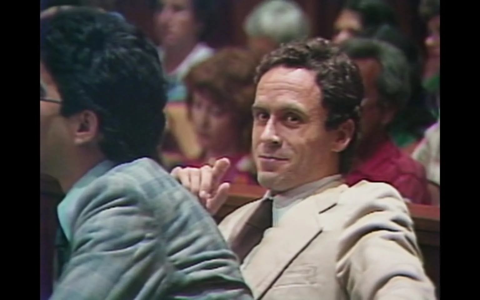 The image depicts a courtroom scene featuring two men seated at a table. The man on the left has dark, curly hair and is wearing a gray suit with a light blue shirt. He appears to be looking down or slightly away from the camera. The man on the right has light brown, wavy hair and is dressed in a light-colored suit, possibly beige, with a white shirt and a dark tie. He is leaning back in his chair, looking directly at the camera with a slight smile and a raised finger, suggesting a gesture of acknowledgment or confidence.

In the background, there are several individuals visible, including a mix of men and women. The audience appears to be engaged, with some looking towards the front of the courtroom. The setting is characterized by wooden paneling and a formal atmosphere typical of a courtroom, with rows of seats filled with spectators.

The lighting in the image is somewhat dim, contributing to the serious tone of the environment. The overall composition captures a moment during a legal proceeding, with a focus on the two men at the forefront, particularly the one with the light suit who is making eye contact with the camera. There are no visible text, signs, or logos in the image. The scene conveys a sense of tension and formality, typical of judicial settings.