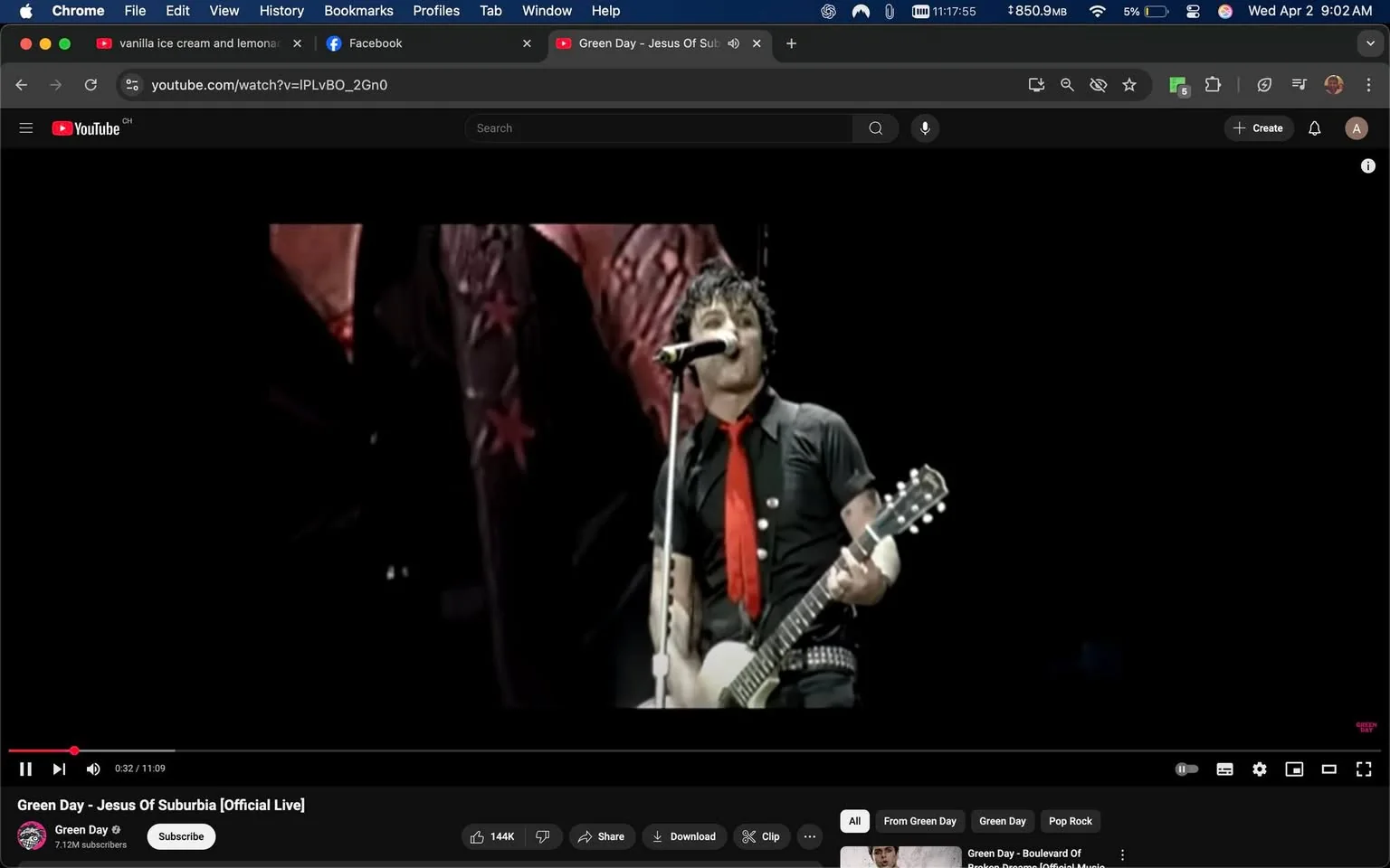 The image depicts a live performance by a musician, likely a member of a rock band. The individual is positioned centrally, holding a white electric guitar with a double cutaway design. The musician has short, dark, tousled hair and is wearing a black short-sleeved shirt with a collar. The shirt is adorned with a few visible buttons and appears to be fitted. A bright red tie is worn loosely around the neck, contrasting with the black shirt. The musician has visible tattoos on both arms, which are partially exposed due to the rolled-up sleeves.

The individual is singing into a microphone, which is mounted on a stand in front of them. Their facial expression appears focused, and they are engaged in the performance. The background features a dark stage setting, with hints of red and black colors, possibly indicating stage lighting or decorations. There are large fabric banners or backdrops with star motifs, adding to the visual elements of the performance space.

In the bottom portion of the image, there are visible elements of a YouTube video interface, including a play bar indicating the video is 32 seconds into an 11-minute and 9-second performance. The title of the video is "Green Day - Jesus Of Suburbia [Official Live]," suggesting that the band performing is Green Day. The channel name "Green Day" is displayed prominently, along with a subscriber count of 7.12 million. The interface includes options to subscribe, share, and download the video, along with a list of related videos.

Overall, the setting is indicative of a live music performance, with a focus on the musician and the energetic atmosphere typical of rock concerts.