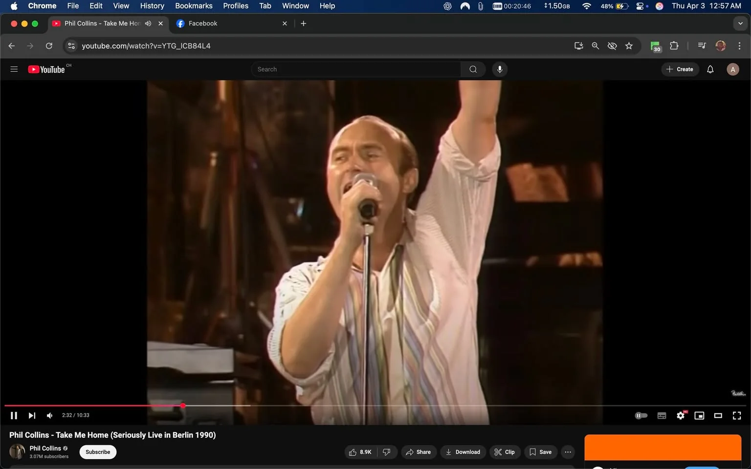 The image shows a live performance scene featuring a male singer. He is positioned centrally, holding a microphone with both hands, and appears to be singing passionately. His right arm is raised above his head, suggesting an energetic or engaging performance. 

The singer has short, light brown hair and is wearing a white shirt with a loose fit. The shirt has a subtle striped pattern, featuring multiple colors, including light blue, pink, and green. The shirt is unbuttoned at the collar, revealing a hint of a darker undershirt beneath. His facial expression is intense, with his mouth open as he sings, and his eyes are focused, likely engaging with the audience.

In the background, there are blurred figures and instruments, indicating a band or musical setup. The lighting is warm and somewhat dim, with a focus on the performer, creating a concert atmosphere. The stage appears to be equipped with various musical instruments, although specific details are not clearly visible due to the focus on the singer.

At the bottom of the image, there is a YouTube video player interface. The title of the video is "Phil Collins - Take Me Home (Seriously Live in Berlin 1990)," indicating that the performance is by Phil Collins. The video has a playtime of 10 minutes and 33 seconds, with a current playback time of 2 minutes and 32 seconds. Below the video, there are options to subscribe, share, download, and clip the video, along with viewer engagement metrics showing 8.9K likes.

The overall setting is a concert stage, likely during the 1990s, given the style of the performance and the video quality. The audience is not visible in this frame, but the context suggests a live music event.