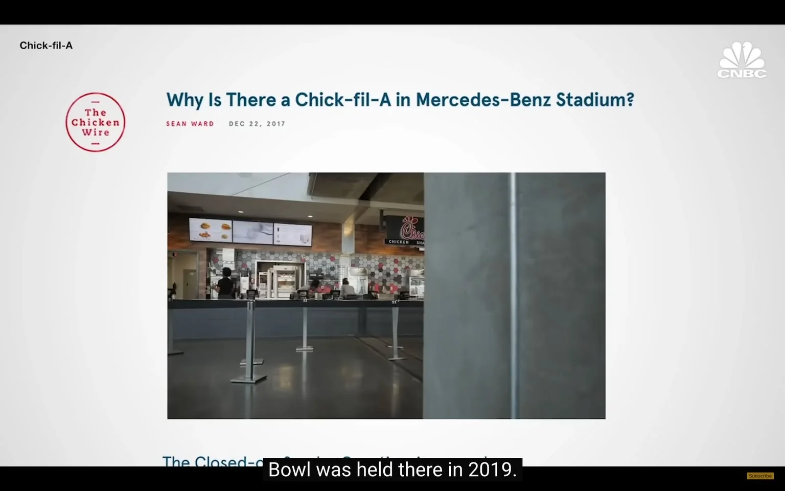 The image features a digital article titled "Why Is There a Chick-fil-A in Mercedes-Benz Stadium?" by Sean Ward, dated December 22, 2017. The article is hosted on a website called "The Chicken Wire." 

In the foreground, there is a view of a Chick-fil-A restaurant located within the stadium. The restaurant's counter is visible, showcasing a menu board with various food items displayed on digital screens above. The menu appears to include chicken sandwiches and other fast-food items, typical of the Chick-fil-A brand. The restaurant's design features a modern aesthetic with a combination of wood and hexagonal tile patterns in the background.

There are several metal stanchions with retractable belts set up in front of the counter, indicating a queue area for customers. The floor is polished and appears to be made of a dark material, likely tile or polished concrete. 

In the background, a few individuals can be seen, but their faces and specific details are not clearly visible. They appear to be engaged in activities related to ordering or preparing food. The overall setting is bright and well-lit, typical of a stadium environment, suggesting a busy atmosphere.

The upper part of the image includes the Chick-fil-A logo, which features a stylized chicken and the brand's name in a distinctive font. The article's title is prominently displayed in a bold, dark blue font, contrasting against a white background. 

No additional text or images are present in the visible area of the image, and the focus remains on the Chick-fil-A restaurant within the stadium context.