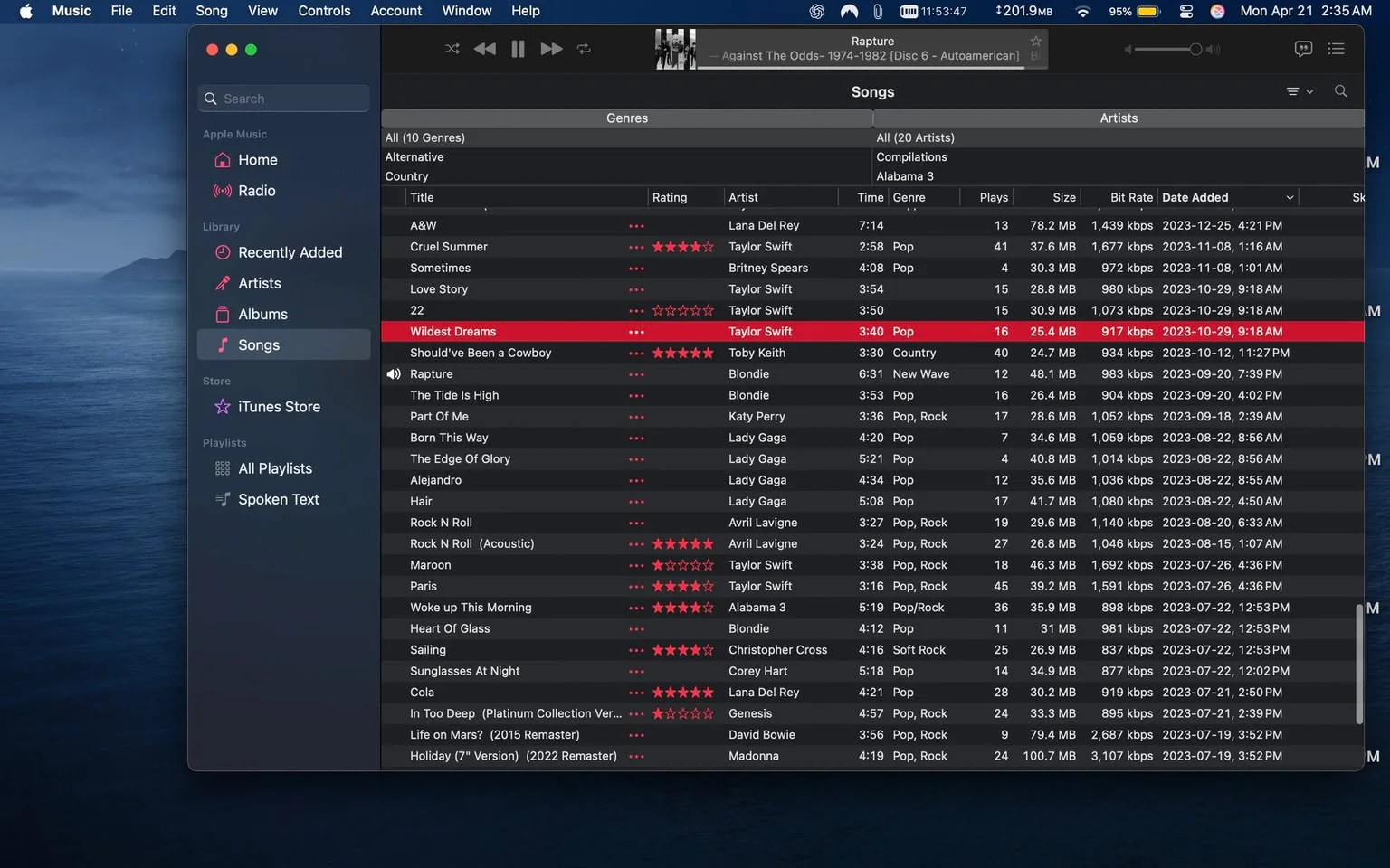 The image displays a screenshot of a music application interface, likely Apple Music, on a Mac computer. The background features a serene ocean view with a mountainous landscape, suggesting a calm environment.

On the left side, there is a sidebar with various sections labeled "Home," "Radio," "Recently Added," "Artists," "Albums," and "Songs." The "Songs" section is highlighted, indicating it is currently selected. Below this, there are options for "Store" and "Playlists," along with a search bar at the top.

The main area of the interface shows a list of songs with several columns: "Title," "Rating," "Artist," "Time," "Genre," "Plays," "Size," "Bit Rate," "Date Added," and "Sk." The songs are organized in a table format, with alternating row colors for better readability. 

Notable entries include:
- "Wildest Dreams" by Taylor Swift, marked in red, indicating it may be currently selected or highlighted. It has a duration of 3:40 minutes and is categorized under the "Pop" genre, with 16 plays and a bit rate of 917 kbps.
- Other songs listed include "Should've Been a Cowboy" by Toby Keith, "Rapture" by Blondie, and "Born This Way" by Katy Perry, among others. Each entry includes details such as the artist, genre, and date added.

The interface also shows star ratings next to some songs, indicating user ratings. The date added for "Wildest Dreams" is noted as October 29, 2023, at 9:18 AM.

At the top of the window, there are playback controls, including play, pause, and skip buttons, along with the title of the album "Against The Odds - 1974-1982 [Disc 6 - Autoamerican]." The time displayed in the top right corner reads 11:54:47 AM.

Overall, the image captures a digital music library interface with a focus on song management and playback features, providing a comprehensive view of the user's music collection.