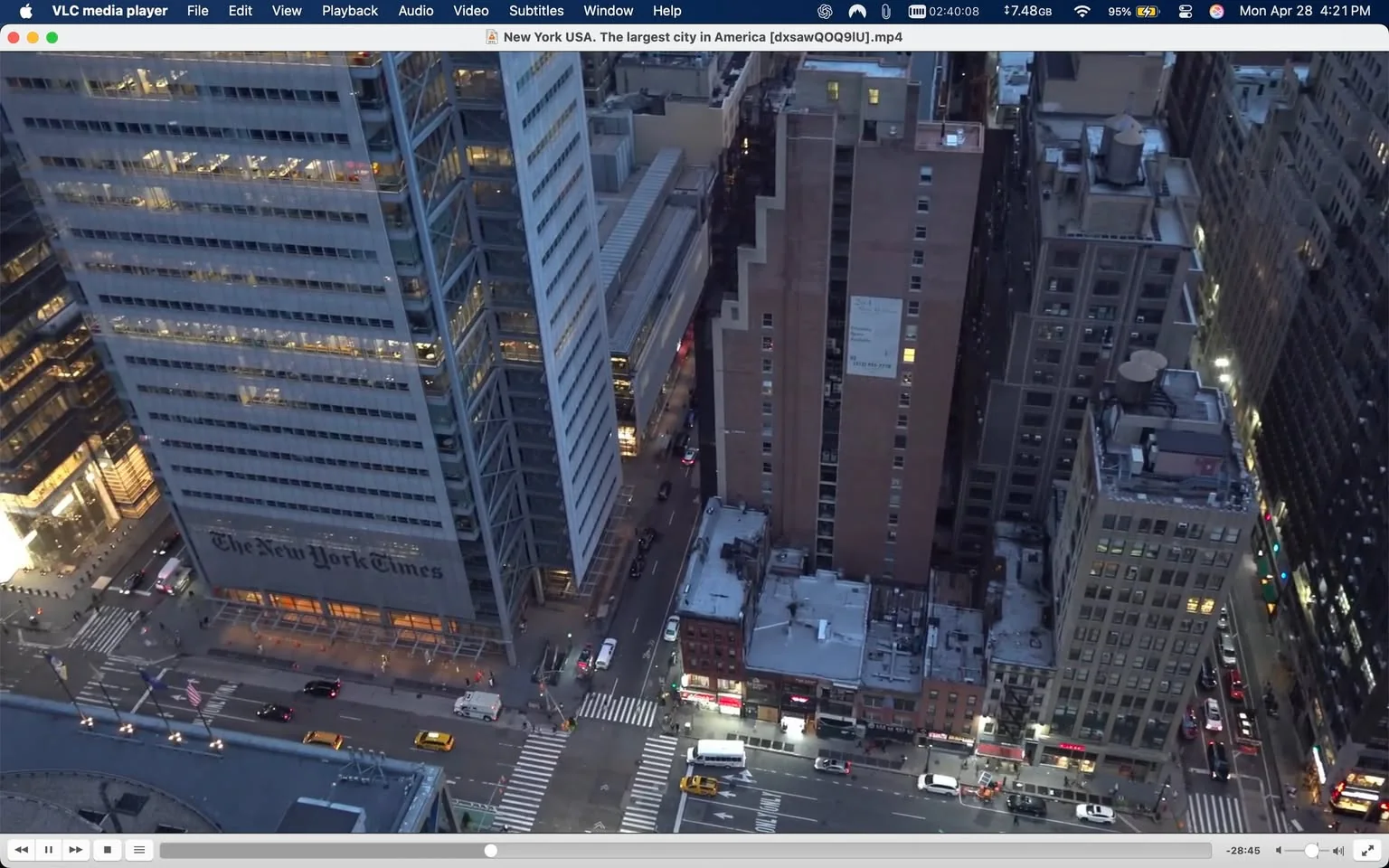 The image depicts a high-angle view of a cityscape, likely in New York City, during twilight. In the foreground, the prominent building features the logo and name of "The New York Times" displayed on its facade, which is a large, modern glass structure. The building has multiple floors with visible office lights on, indicating activity inside.

To the right of this building, there is a taller, brick structure with a stepped design on its side. The rooftop of this building shows various mechanical units and a water tower. The surrounding buildings are a mix of modern glass and older brick architecture, showcasing the urban landscape's diversity.

The streets below are busy with vehicles, including yellow taxis and other cars, moving along the avenues. There are crosswalks visible, with pedestrians appearing small in comparison to the towering buildings. Streetlights are illuminated, casting a warm glow on the streets.

In the background, the skyline features a variety of building heights and styles, contributing to the dense urban environment. The sky is transitioning from blue to darker shades, indicating the approach of night. The overall atmosphere suggests a bustling city life, with a blend of work and leisure activities occurring simultaneously.

No people are clearly visible in the image, and there are no discernible facial expressions or clothing details to describe. The focus remains on the architectural elements and the urban setting.