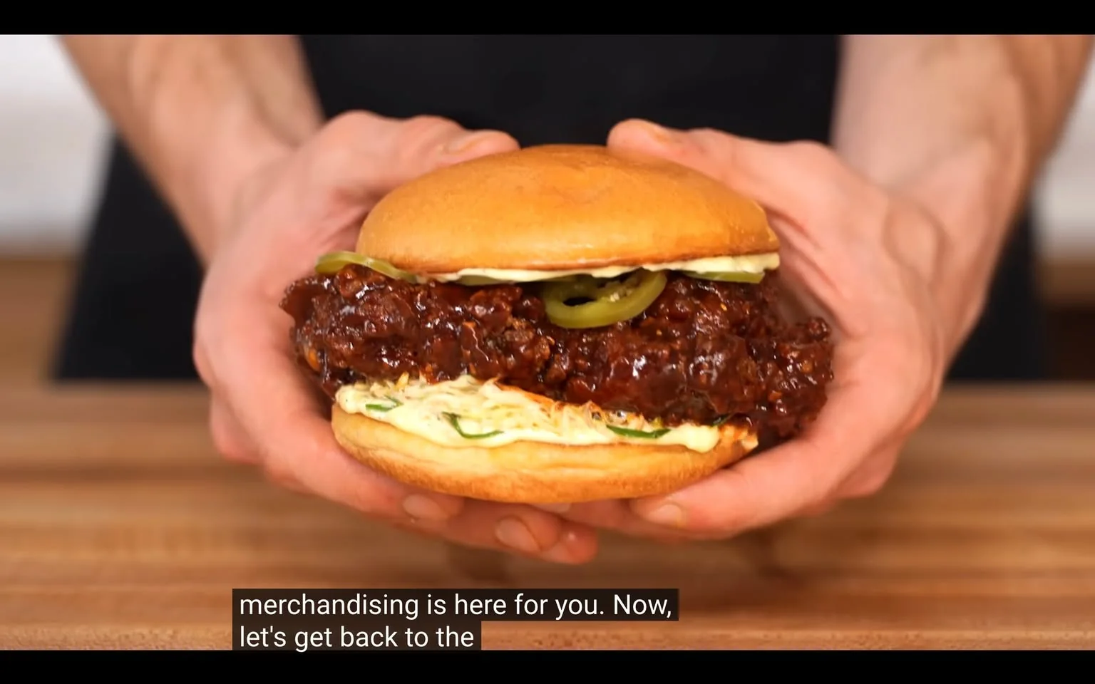The image features a close-up of a sandwich being held by a pair of hands. The sandwich consists of a soft, golden-brown bun that appears slightly toasted on the top. The bottom half of the bun is visible, revealing a generous portion of dark, glossy sauce-covered meat, likely fried chicken, which is thick and has a rich, sticky texture. 

On top of the meat, there are two slices of green jalapeño peppers, adding a pop of color and suggesting a spicy flavor. Below the meat, there is a layer of shredded lettuce, which is pale in color and adds a fresh element to the sandwich. A creamy sauce, possibly mayonnaise or a similar condiment, is spread on the inside of the top bun, enhancing the overall flavor profile.

The hands holding the sandwich are partially visible, with the fingers wrapped around the sides of the bun. The background is blurred, but it appears to be a wooden surface, likely a countertop or table, which suggests a casual cooking or dining setting. The lighting is bright, highlighting the textures and colors of the sandwich, making it look appetizing.

There are no visible logos, trademarks, or readable text in the image. The focus is entirely on the sandwich and the hands holding it, with no identifiable people or brands present. The overall composition emphasizes the food item, making it the central subject of the image.