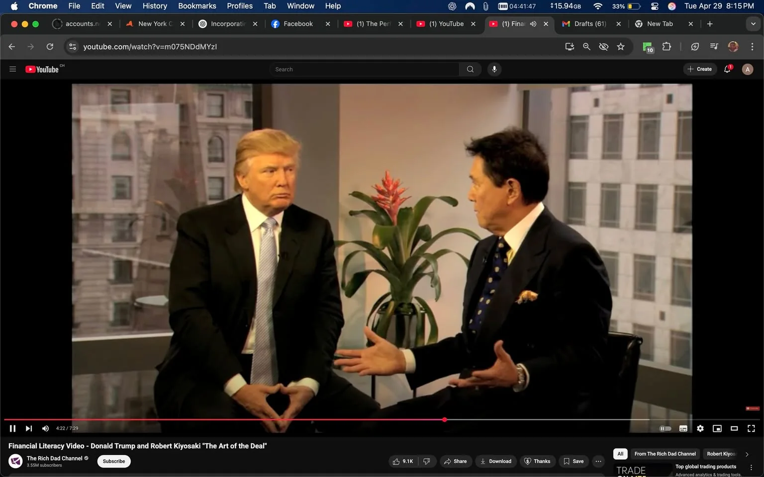 The image shows two men seated in a well-lit indoor setting, likely an office or studio. The background features large windows with a view of a cityscape, including buildings and a hint of greenery. 

On the left, a man with light brown hair styled in a comb-over is wearing a black suit with a white dress shirt and a light blue tie. His expression is serious, and he is sitting with his hands clasped in front of him, fingers interlocked. 

On the right, another man with dark hair, slightly graying at the temples, is dressed in a dark suit with a white shirt and a patterned tie featuring blue and gold dots. He appears to be animatedly speaking, gesturing with his right hand while his left hand rests on his lap. 

In the background, there is a potted plant with long green leaves and a red flower, adding a touch of color to the scene. The overall atmosphere is professional, and the men seem to be engaged in a serious conversation. 

There are no visible logos, trademarks, or text in the image, and the focus is primarily on the two individuals and their interaction. The lighting is bright, enhancing the clarity of the scene.