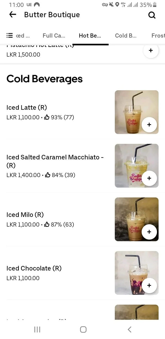 The image displays a digital menu from a café or restaurant named "Butter Boutique." The top of the menu shows the name "Butter Boutique" prominently, likely in a stylized font. The menu is divided into sections, with the current section labeled "Cold Beverages."

Under the "Cold Beverages" section, there are several items listed, each with specific details:

1. **Iced Latte (R)**:
   - Price: LKR 1,100.00
   - Rating: 93% (77 reviews)
   - Accompanied by an image of the drink in a clear cup with a black straw, featuring the "Butter Boutique" logo.

2. **Iced Salted Caramel Macchiato - (R)**:
   - Price: LKR 1,400.00
   - Rating: 84% (39 reviews)
   - The drink is shown in a clear cup, similar to the Iced Latte, with a light-colored beverage.

3. **Iced Milo (R)**:
   - Price: LKR 1,100.00
   - Rating: 87% (63 reviews)
   - The image shows a drink in a clear cup, with a darker beverage.

4. **Iced Chocolate (R)**:
   - Price: LKR 1,100.00
   - The image features a drink in a clear cup with a layered appearance, possibly indicating chocolate and cream.

Each item includes a "+" symbol, likely indicating an option to add the item to an order. The background of the images appears to be a neutral, possibly textured surface, which does not distract from the beverages.

The overall layout is clean and organized, with a focus on readability and visual appeal. The menu does not include any people, clothing, or facial expressions, as it is solely focused on the beverage offerings. There are no additional environmental context indicators, such as location or setting, beyond the café branding.