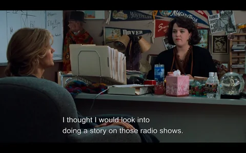 The image depicts a scene in an office setting. There are two women engaged in conversation. 

1. **People**:
   - **Woman on the left**: She has long, wavy hair and is wearing a dark gray sweater. She is seated in a chair, facing the woman on the right. Her expression appears thoughtful as she speaks.
   - **Woman on the right**: She has short, curly dark hair and is wearing a black cardigan over a brown top. She is seated at a desk, looking directly at the woman on the left with a neutral expression.

2. **Desk and Surroundings**:
   - The desk is cluttered with various items:
     - A stack of file folders is visible, organized in a metal holder.
     - There are several drinks, including a blue cup and a water bottle.
     - A pink box, possibly containing tissues, is on the desk, along with a small glass container filled with colorful candies.
     - A snow globe is also present on the desk.
   - Behind the woman on the right, there are various decorations and items on the wall, including:
     - Banners and signs with names of places like "Montpelier" and "Dover."
     - A mannequin dressed in a colorful cape and a hat, positioned in the background.
     - A whiteboard with handwritten notes and diagrams, including the words "Add" and "Ad."

3. **Setting**:
   - The office appears to be a casual workspace, possibly related to media or journalism, given the context of the conversation about radio shows.
   - The walls are adorned with various memorabilia and posters, contributing to a creative atmosphere.

4. **Text**:
   - There is visible text at the bottom of the image, which reads: "I thought I would look into doing a story on those radio shows."

Overall, the scene captures a moment of dialogue in a creative office environment, with various personal and professional items visible in the setting.