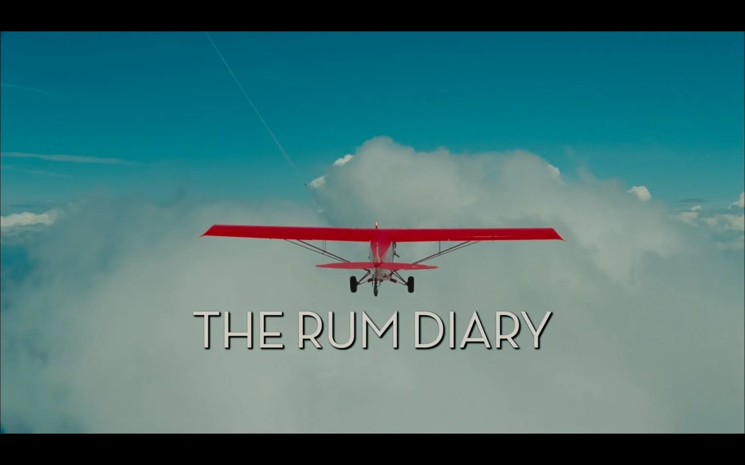 The image features a small red airplane flying above a thick layer of white clouds against a bright blue sky. The airplane is seen from a rear angle, showcasing its distinctive red wings and tail. The propeller is visible at the front of the aircraft, and the landing gear consists of two small wheels. The background is predominantly sky blue with a few wispy clouds, creating a serene and expansive atmosphere.

At the bottom of the image, the title "THE RUM DIARY" is prominently displayed in large, bold, white letters. The font is simple and modern, contrasting well with the vibrant colors of the airplane and the sky. The text is centered and spans the width of the image, providing a clear focal point.

There are no visible people, objects, or additional text in the image apart from the airplane and the title. The overall composition conveys a sense of adventure and exploration, typical of aerial imagery. The absence of any logos, trademarks, or identifiable brands beyond the title suggests that this is likely a promotional image for a film or media project. The setting appears to be high in the atmosphere, emphasizing the airplane's flight above the clouds.