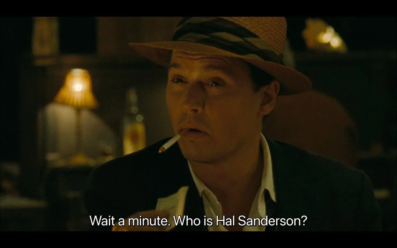 The image features a man seated at a table in a dimly lit setting, likely a bar or restaurant. He has a cigarette in his mouth and is wearing a straw hat with a dark band around it. His attire includes a dark jacket over a white shirt, which is partially unbuttoned at the collar. The man has a slightly furrowed brow and a contemplative expression, suggesting he is engaged in thought or conversation.

In the background, there are warm, ambient lights, including a lamp with a pleated shade that casts a soft glow. A bottle is visible on the table behind him, although the label is not clearly discernible. The overall atmosphere appears relaxed and intimate, typical of a social setting.

The text overlay at the bottom reads: "Wait a minute. Who is Hal Sanderson?" This indicates that the man is likely involved in a dialogue or narrative context, but no additional context about the conversation is provided in the image.

The background is somewhat blurred, focusing attention on the man and his expression. The color palette is warm, with browns and soft yellows dominating the scene, contributing to the cozy ambiance. The setting does not provide any specific indicators of time or place beyond the general bar or restaurant environment.