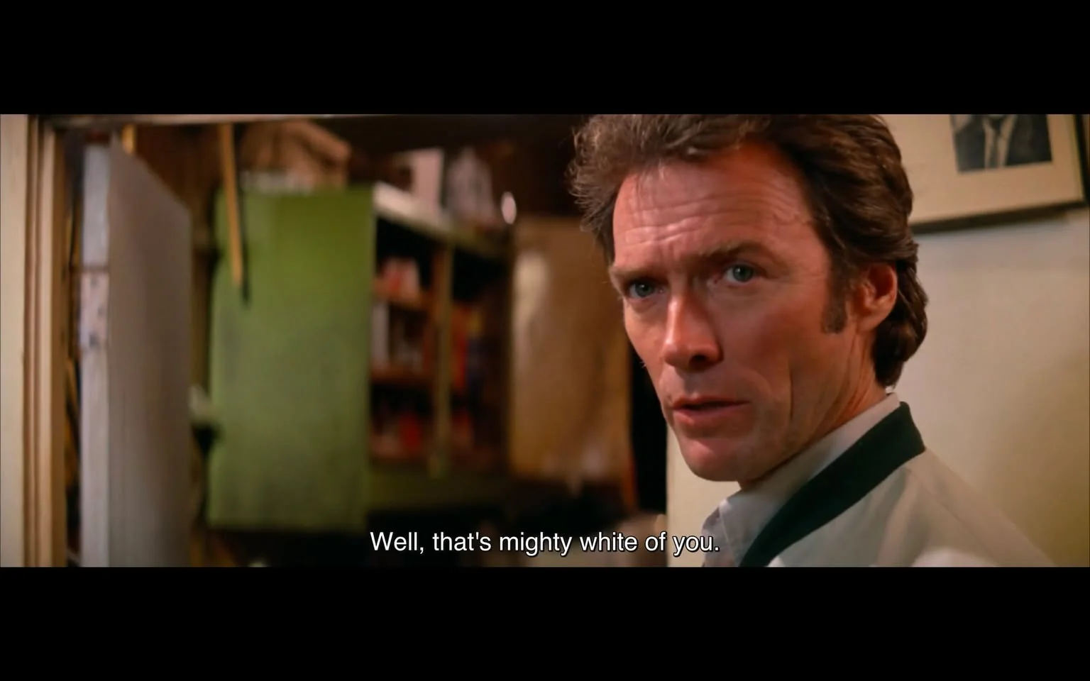 The image depicts a scene from a film featuring a male character with a serious expression. He has short, slightly tousled hair and is wearing a white shirt with a dark collar. The character's face is partially turned towards the camera, revealing a focused gaze and a hint of tension in his expression. 

In the background, there is a green object that appears to be a cabinet or a shelf, suggesting a cluttered indoor environment. The setting has a somewhat rustic or vintage feel, with shelves visible that contain various items, though the details of those items are not clearly discernible. The lighting in the scene is warm, contributing to the overall atmosphere.

The visible text at the bottom of the image reads: "Well, that's mighty white of you." This line suggests a dialogue exchange, but the context of the conversation is not provided in the image. 

Overall, the scene conveys a moment of intensity, with the character's expression and the surrounding environment hinting at a deeper narrative, though specific details about the plot or characters are not available from this image alone.