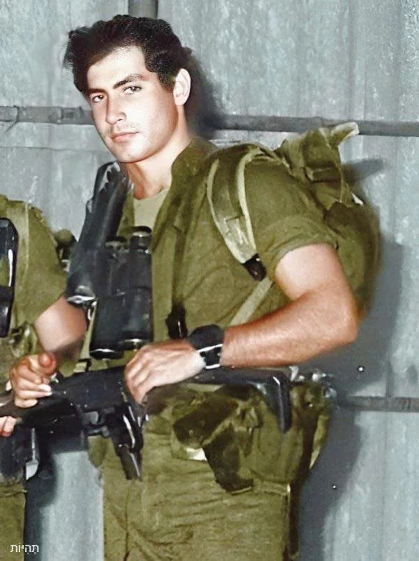 The image depicts a young man in military attire, standing in a relaxed pose. He is wearing a green military uniform, which consists of a short-sleeved shirt and cargo pants. The shirt is fitted, and the sleeves are rolled up to the elbows, revealing his forearms. He has a tactical vest or harness over his shirt, which is equipped with various pouches. The man is holding a firearm in his right hand, with his left hand resting on the weapon. 

He has dark hair, styled in a slightly wavy manner, and his facial expression appears serious yet composed. The background is a metallic surface, possibly a wall or a vehicle, which is gray and textured. There are two other individuals partially visible in the background, also dressed in similar military attire, but their faces are not clearly visible.

The image has been colorized, giving it a vibrant appearance, with the green of the uniform standing out against the gray backdrop. There are no visible logos, text, or identifiable brands in the image. The overall setting suggests a military context, possibly a training facility or deployment area. The lighting appears natural, highlighting the subjects and their attire.