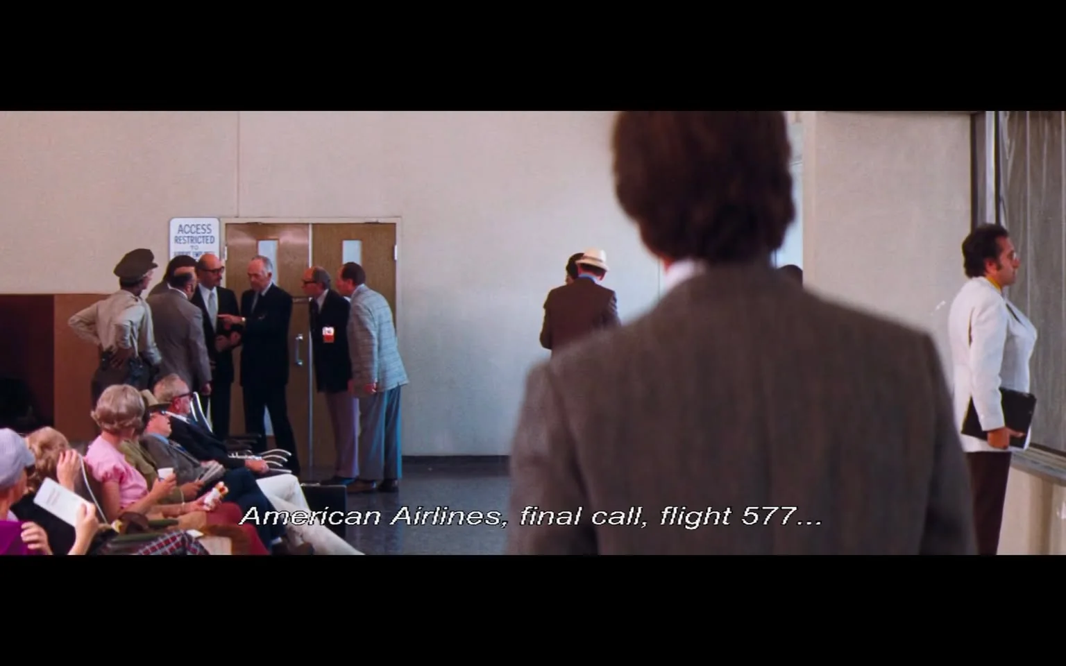 The image depicts a scene in an airport terminal. In the foreground, a man in a gray suit stands with his back to the camera, partially obscuring his face. He appears to be looking towards a group of people gathered near a set of double doors marked with a sign that reads "ACCESS RESTRICTED." 

The group consists of several men in suits, engaged in conversation. One man, wearing glasses and a dark suit, is pointing towards another individual, who is dressed in a light blue blazer. The other men appear to be listening intently, with expressions of concern or interest. A police officer in a tan uniform stands to the left of the group, with his hands on his hips, observing the situation.

In the background, a few passengers are seated in the waiting area. They are dressed in a mix of casual and semi-formal attire, including a woman in a pink blouse and a man in a plaid suit. Some individuals are holding papers or magazines, while others appear to be engaged in conversation. The overall atmosphere suggests a busy airport environment.

The text at the bottom of the image reads: "American Airlines, final call, flight 577..." indicating an announcement related to a flight. The walls of the terminal are painted a light color, and the flooring appears to be a dark tile, contributing to the typical airport setting. The lighting is bright, typical of an indoor terminal space.