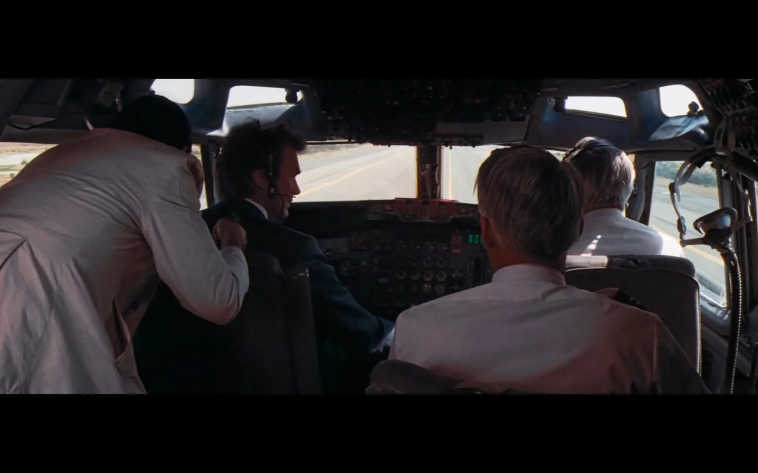 The image depicts the cockpit of an aircraft, showing a view from behind the pilots. There are three men visible, all dressed in formal attire. 

1. **Pilot in the Center**: The central figure, presumably the pilot, is wearing a dark suit and a headset. He is focused on the controls in front of him, which include various buttons, switches, and screens. The cockpit is equipped with numerous instruments, including gauges and lights, indicating the aircraft's status.

2. **Co-Pilot**: To the right of the pilot is another man, also in a white shirt, who appears to be the co-pilot. He is seated and looking towards the front, possibly monitoring the flight instruments or assisting the pilot.

3. **Third Person**: To the left, a third man in a light-colored suit is leaning over the pilot's shoulder. His expression is not clearly visible, but he seems to be engaged in the situation, possibly providing assistance or guidance.

4. **Cockpit Details**: The cockpit features a large array of controls, including a control yoke, throttle levers, and various displays. The background shows a clear view of the runway ahead, with the ground visible through the cockpit windows. The lighting suggests it is daytime, with natural light illuminating the cockpit.

5. **Setting**: The environment appears to be an airport setting, with the runway visible through the front windows. The overall atmosphere is focused and professional, typical of an aircraft cockpit during takeoff or landing preparations.

No readable text, logos, or trademarks are visible in the image. The focus remains on the individuals and the cockpit instruments.