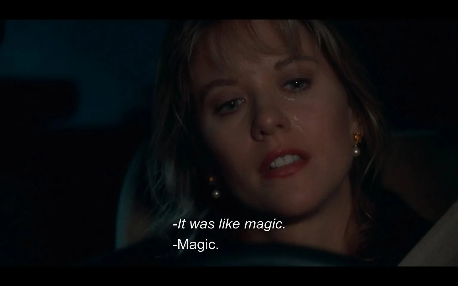 The image depicts a close-up of a woman sitting in a car, with a dark background suggesting nighttime. She has light brown hair that appears slightly wavy and is styled with some bangs. Her facial expression shows a mix of contemplation and emotion, with her eyes slightly glistening, possibly indicating tears. She has a subtle sheen on her cheeks, which may suggest she has been crying or is emotional. 

The woman is wearing small, round pearl earrings that stand out against her skin. Her lips are painted a soft shade of red, and her makeup is understated, enhancing her natural features. She is dressed in a dark-colored top, which is not fully visible due to the angle of the shot.

In the foreground, part of the car's interior is visible, including a section of the steering wheel and the dashboard, which is mostly out of focus. The lighting in the car is dim, with a blueish tint that adds to the nighttime atmosphere. 

Text is visible at the bottom of the image, indicating dialogue: "-It was like magic." and "-Magic." The text is white and clearly legible against the darker background. 

The overall setting suggests a private moment, likely a conversation taking place in the car, but no specific location or external environment is visible beyond the interior of the vehicle. There are no identifiable brands, logos, or trademarks present in the image.