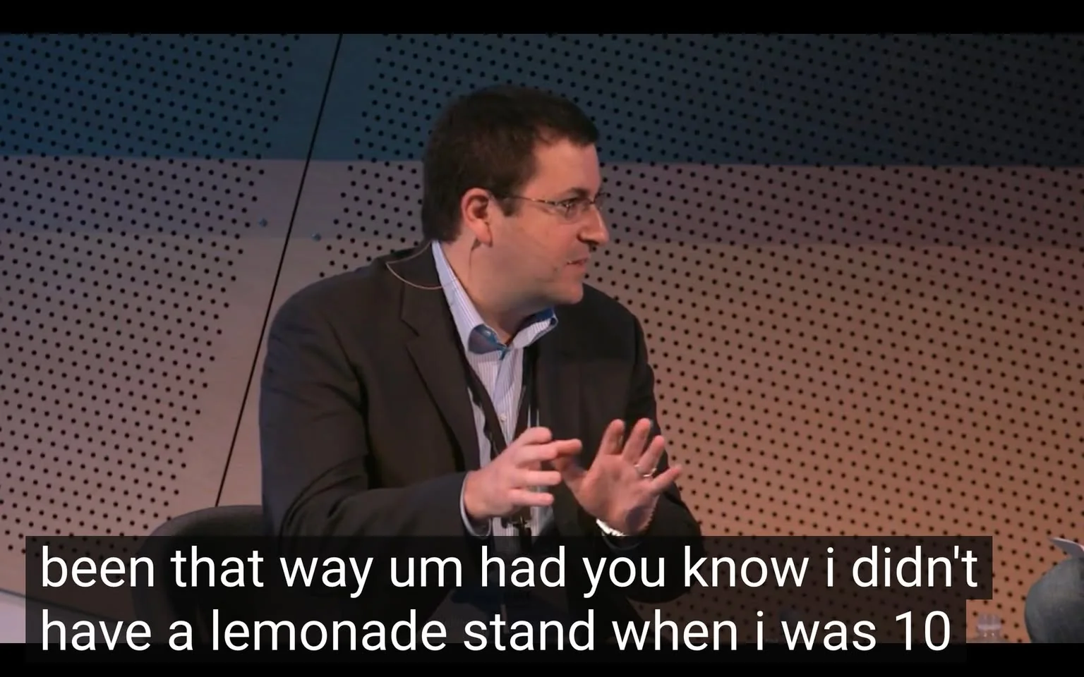 The image shows a man speaking at a conference or panel discussion. He has short, dark hair and is wearing glasses. His attire consists of a dark blazer over a light blue dress shirt, which is unbuttoned at the collar. He appears to be animated in his speech, using hand gestures to emphasize his points. 

The background features a textured wall with a pattern of small holes, suggesting a modern venue designed for acoustics. The lighting is bright, illuminating the speaker's face and the area around him. 

In front of the speaker, there is a microphone attached to his collar, indicating that he is likely addressing an audience. A water bottle is visible on the table in front of him, but no other objects or people are clearly discernible in the immediate vicinity. 

The text overlay at the bottom of the image includes a transcription of what he is saying, which mentions not having a lemonade stand when he was 10 years old. The overall setting appears to be professional, likely part of a larger event focused on discussions or presentations.
