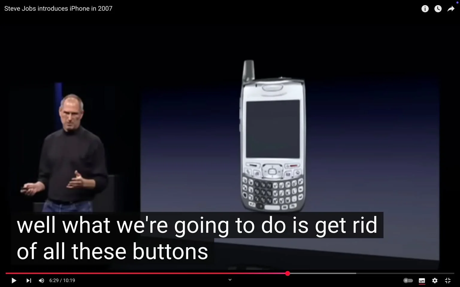 The image depicts a presentation scene featuring a man, presumably Steve Jobs, standing on a stage. He is wearing a black turtleneck sweater and blue jeans. His expression appears focused as he gestures with his right hand while holding a small device in his left hand. The background is dark, with a gradient transitioning from black to blue, creating a professional atmosphere.

To the right of the speaker, there is a white mobile phone displayed on a stand. The phone has a rectangular shape with a large screen and a physical keyboard below it, featuring numerous buttons. The device appears to have a small antenna on the top, indicating it is an older model. The overall design of the phone suggests it is from the early 2000s, characterized by its bulkiness and the presence of a QWERTY keyboard.

The text overlay at the bottom of the image reads: "well what we're going to do is get rid of all these buttons," indicating that the speaker is discussing the transition from traditional mobile phones with physical buttons to a more modern design, likely referencing the introduction of the iPhone.

The setting appears to be a tech conference or product launch event, given the formal presentation style and the presence of a large audience, although the audience is not visible in this particular frame. The overall tone of the image suggests a significant moment in technology history, likely related to the unveiling of a groundbreaking product.