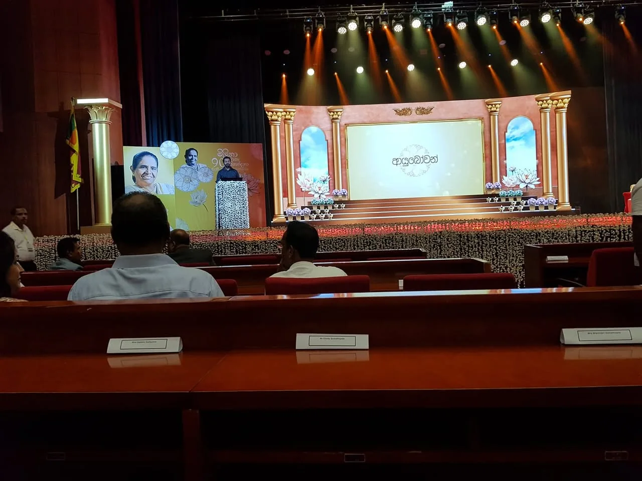 The image depicts an indoor event space, likely a stage set up for a formal gathering or celebration. The stage is adorned with elaborate decorations, featuring large, ornate columns on either side, which are painted in a light color. The backdrop includes a large screen displaying text in a decorative font, possibly in a South Asian language, with floral motifs surrounding it. 

To the left of the stage, there are two large portraits of individuals, one of whom appears to be a woman, smiling, while the other is a man, both possibly significant figures in the context of the event. The portraits are set against a bright yellow background, enhancing their visibility.

The seating area in the foreground consists of red upholstered chairs arranged in rows. There are several people seated, some facing the stage, while others are engaged in conversation. The individuals are dressed in formal attire; one man is wearing a light blue shirt, while another is in a white shirt. The audience appears to be a mix of men and women, with a few visible in the frame.

In the background, there is a decorative arrangement of flowers or fabric that covers the floor, adding to the festive atmosphere. The lighting is bright, with multiple spotlights illuminating the stage, creating a vibrant ambiance.

A Sri Lankan flag is visible to the left side of the image, indicating the location or cultural significance of the event. The overall setting suggests a celebratory occasion, possibly a cultural or commemorative event, given the formal decorations and attire of the attendees.