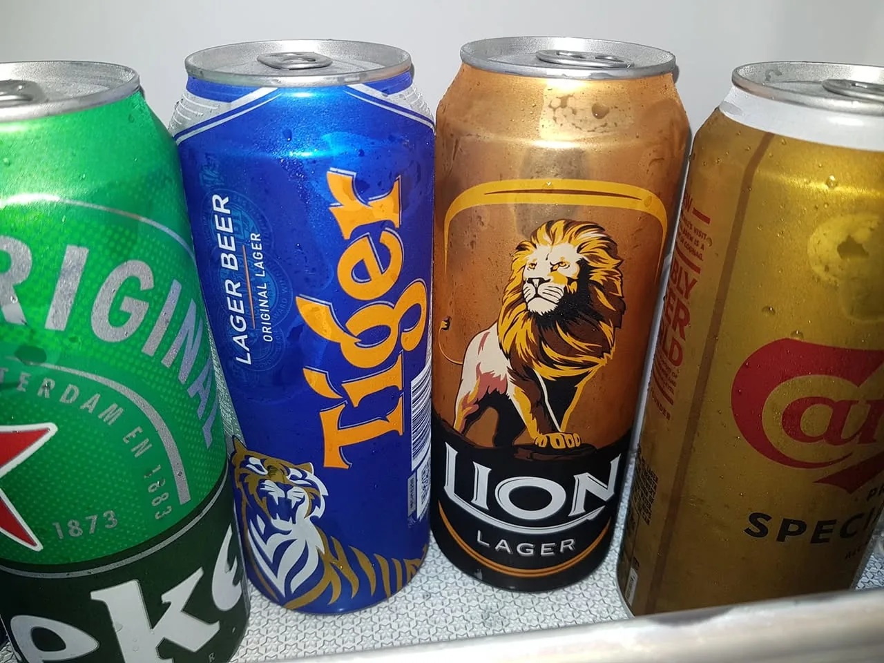 The image shows a selection of beer cans arranged closely together, likely inside a refrigerator. 

1. **Cans and Labels**:
   - **Leftmost Can**: Green in color with white text that reads "Heineken" prominently displayed. The label includes the word "ORIGINAL" and "AMSTERDAM" along with the year "1873."
   - **Second Can**: Blue with orange text that reads "Tiger" and features a graphic of a tiger. The label states "LAGER BEER" and "ORIGINAL LAGER."
   - **Third Can**: Gold in color with a prominent illustration of a lion, labeled "Lion Lager." The lion has a detailed mane and a fierce expression, and the can is designed to evoke a sense of strength.
   - **Rightmost Can**: Yellow with red text that reads "Carlsberg" and "SPECIAL." The design is simple, with a focus on the brand name.

2. **Visual Features**:
   - The cans are metallic and shiny, with condensation visible on their surfaces, indicating they are cold.
   - The arrangement of the cans is slightly overlapping, with the labels facing outward for visibility.

3. **Background and Setting**:
   - The background is a light-colored surface, possibly the interior of a refrigerator, with a textured pattern that suggests a cooling element.
   - The lighting appears bright, highlighting the colors and condensation on the cans.

Overall, the image captures a selection of popular beer brands, showcasing their distinctive designs and colors in a chilled environment.