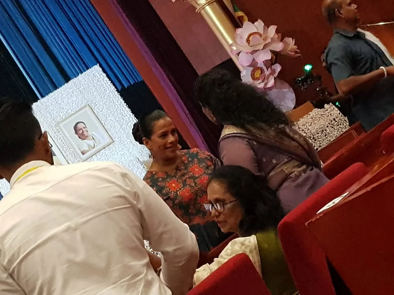 The image depicts a gathering in a formal setting, likely an event or ceremony. The background features a stage with a blue curtain and a decorative white panel that has a framed photograph displayed on it. The photograph appears to show a person, but the details are not discernible.

In the foreground, several individuals are engaged in conversation. On the left, a man is partially visible, wearing a white shirt with the collar turned down and a yellow lanyard around his neck. He has short, dark hair and is facing away from the camera.

To the right, a woman with dark hair styled in a bun is smiling and wearing a floral-patterned dress with shades of red, orange, and green. She appears to be interacting with another woman who has long, curly hair and is dressed in a purple outfit with a decorative shawl. This second woman is facing the first and seems to be engaged in conversation.

In the bottom right corner, another woman with glasses is seated, wearing a white top with a greenish overlay. She appears to be looking at something, possibly engaged in a discussion with the others.

The seating arrangement consists of red chairs, and the overall ambiance suggests a formal or celebratory event. There are additional decorations, including large paper flowers, which add to the festive atmosphere. The lighting appears bright, indicating a well-lit indoor environment. 

Overall, the image captures a moment of social interaction among attendees at a formal gathering, with a focus on their clothing and the event's decor.