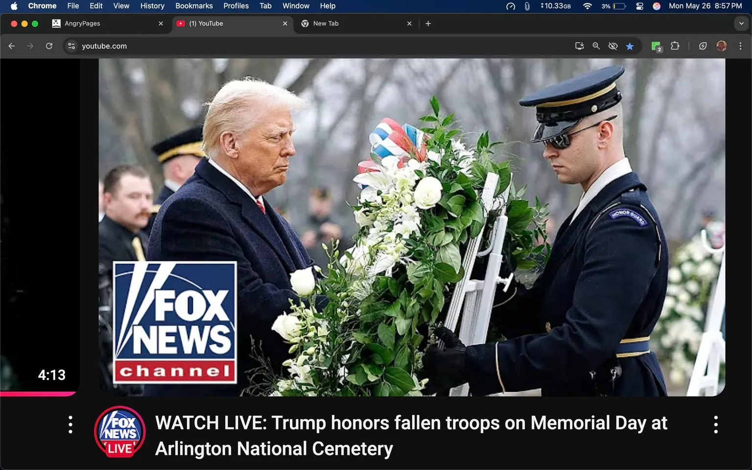 The image depicts a scene at a memorial event, likely at Arlington National Cemetery. In the foreground, a man with light-colored hair, wearing a dark coat, is positioned on the left side of the image. He appears to be solemn and is looking down at a floral arrangement. The arrangement consists of white flowers and green foliage, with some red, white, and blue ribbons visible among the greenery.

To the right of this man, a soldier in a formal military uniform is holding the floral arrangement. The soldier is wearing a dark jacket with a white shirt underneath and a black cap. The jacket has a patch on the shoulder that reads "HONOR GUARD." The soldier's expression is serious, and he is focused on the task at hand, which appears to be presenting the floral tribute.

In the background, several individuals in military attire can be seen, indicating that this is a formal ceremony. The setting appears to be outdoors, with trees in the background that are bare, suggesting it might be a cool or chilly day. The overall atmosphere is respectful and somber, typical of memorial events.

At the bottom left corner of the image, there is a logo for "FOX NEWS CHANNEL," which is prominently displayed in blue and white. Above this logo, there is a text overlay that reads, "WATCH LIVE: Trump honors fallen troops on Memorial Day at Arlington National Cemetery." The text is in white against a black background, indicating that this image is likely part of a live broadcast or news coverage.

The image is framed within a browser window, with tabs and a menu bar visible at the top, indicating that it is being viewed on a computer. The time displayed in the top right corner shows "Mon May 26 8:57 PM," suggesting the date and time of the event or the moment the image was captured.