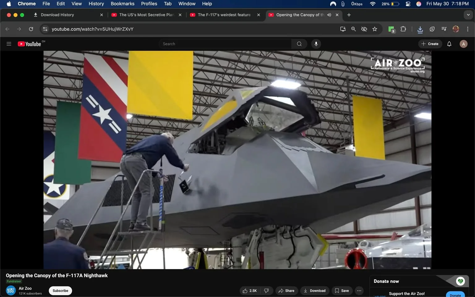 The image depicts a scene inside a hangar where an F-117A Nighthawk aircraft is being worked on. The aircraft is primarily gray with angular shapes and a stealthy design, characteristic of its military purpose. The cockpit area is partially open, revealing some internal components. 

In the foreground, a person is standing on a ladder, leaning slightly forward while using a tool to work on the aircraft. The individual is wearing a blue long-sleeve shirt and gray pants, with their hair partially visible. Their expression is not clearly visible due to the angle. 

In the background, another person, dressed in a dark jacket and a cap, is observing the work being done. The hangar features a high ceiling with visible metal beams and several large banners hanging from it. The banners include a red and blue design with a star, and a yellow banner, among others, indicating a military or aviation theme.

The floor of the hangar is concrete, and various tools and equipment can be seen nearby, suggesting an active maintenance or restoration environment. The lighting is bright, illuminating the aircraft and the workers. The overall atmosphere is one of focused activity, centered around the maintenance of the F-117A Nighthawk.