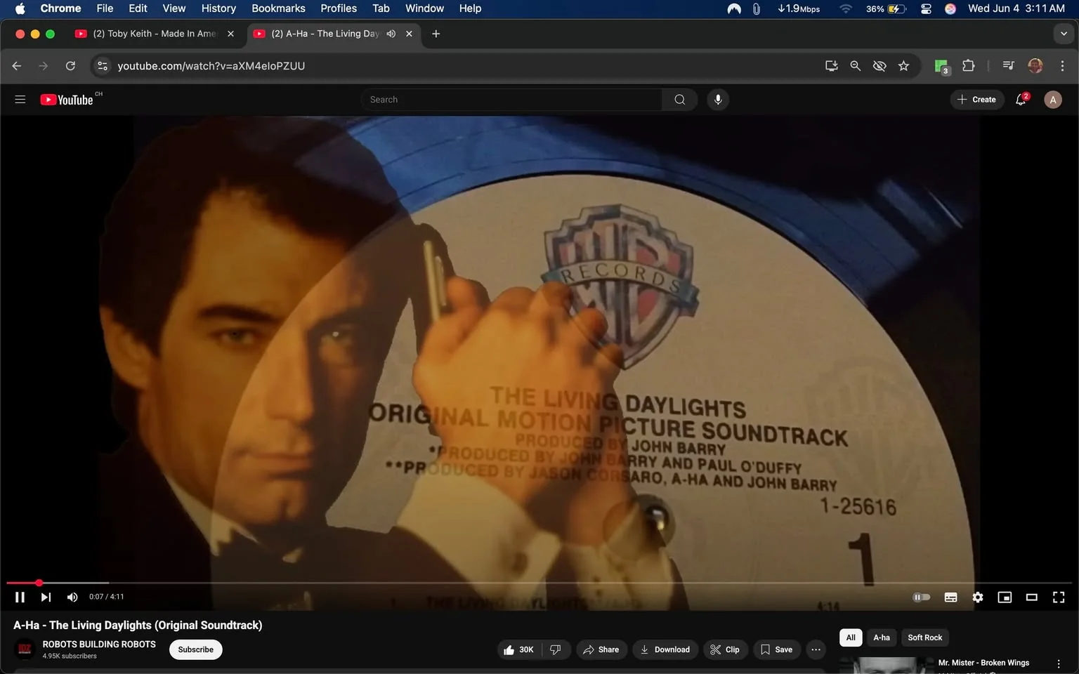 The image features a close-up view of a vinyl record, specifically the soundtrack for "The Living Daylights." The record label is prominently displayed, indicating it is a Warner Bros. Records release. The label includes the title "THE LIVING DAYLIGHTS" in bold text, along with the phrase "ORIGINAL MOTION PICTURE SOUNDTRACK." Below this, it mentions the producers: "PRODUCED BY JOHN BARRY AND PAUL O'DUFFY" and "PRODUCED BY JASON CORSARO, A-HA AND JOHN BARRY." The catalog number "1-25616" is also visible.

In the foreground, there is an image of a man holding a gun to his head, suggesting a tense or dramatic pose. His face is partially obscured, but he has dark hair and is dressed in a formal suit, likely a tuxedo, with a white shirt and black bow tie. The expression on his face appears serious, and he is looking directly at the viewer. The background is blurred, focusing attention on the record and the man's image.

The overall color scheme includes dark tones, with the record label being predominantly white with blue accents. The image has a cinematic quality, likely referencing the film's themes of espionage and action. The setting appears to be a digital interface, as indicated by the YouTube player controls visible at the bottom of the image, including play, pause, and volume options. The video title "A-Ha - The Living Daylights (Original Soundtrack)" is also displayed, indicating the content of the video. 

No additional identifiable brands or logos are present outside of the Warner Bros. Records label. The image does not provide any further context regarding the location or event.
