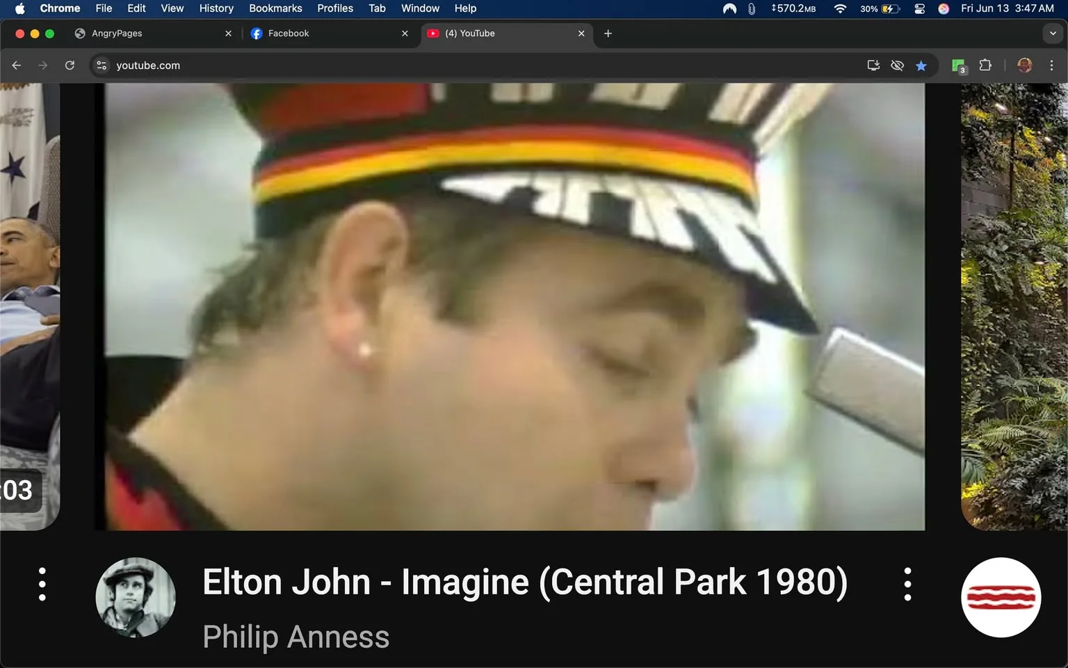 The image appears to be a still from a video featuring a male performer, possibly Elton John, as indicated by the text overlay. He is wearing a distinctive hat that has a black and red color scheme, with a wide brim and a design that includes vertical stripes. The hat has a band of red, yellow, and black around it. The performer has short, light-colored hair that is slightly tousled, and he is wearing a black jacket with red accents. 

His left ear is visible, adorned with a small earring. The performer is positioned in profile, with his eyes closed, suggesting he is either singing or deeply focused on his performance. The microphone is positioned close to his mouth, indicating he is likely in the midst of a live performance.

In the background, there is a blurred setting that appears to be an outdoor venue, possibly a park, given the context of the video title "Imagine (Central Park 1980)." The background is not detailed, but it suggests a crowd or audience, as indicated by the soft focus.

The lower part of the image contains text that reads "Elton John - Imagine (Central Park 1980)" along with the name "Philip Anness" below it. The text is in white against a black background, providing a clear contrast. There are also icons for YouTube and other social media platforms visible in the top browser bar, indicating that this image is part of a video interface.

Overall, the image captures a moment of performance, highlighting the artist's attire and the setting, while providing contextual information through the text overlay.