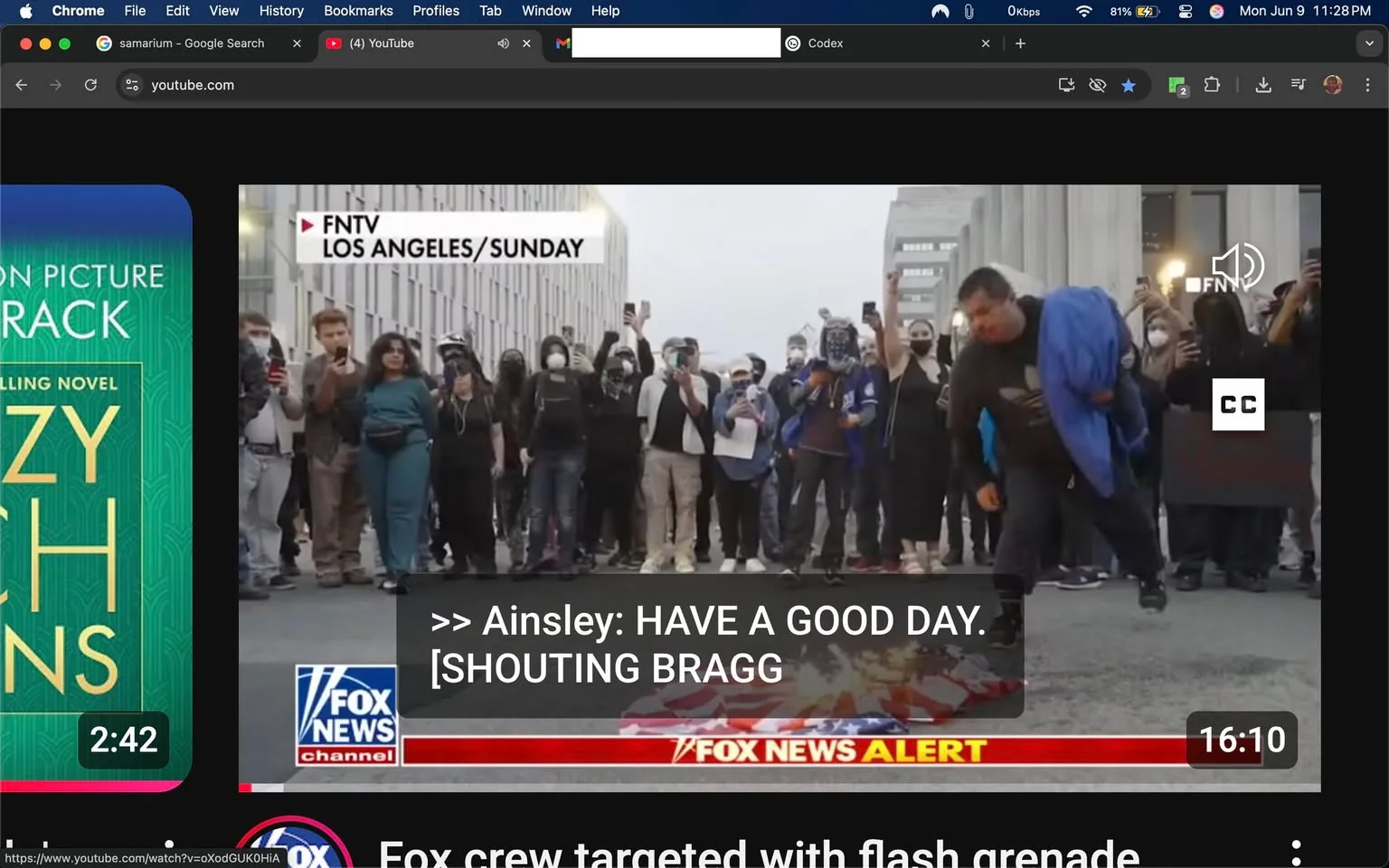 The image depicts a scene from a protest or demonstration taking place in an urban environment, likely in Los Angeles, as indicated by the text in the upper left corner. 

**Text and Logos:**
- The top left corner features the text "FNTV LOS ANGELES/SUNDAY."
- There is a banner at the bottom with the Fox News logo, stating "FOX NEWS ALERT" and additional text that reads "Fox crew targeted with flash grenade."
- A caption appears in the middle of the image, which includes the name "Ainsley" followed by "HAVE A GOOD DAY." and "[SHOUTING BRAGG]."
- The video duration is shown as "16:10" in the bottom right corner.

**People and Clothing:**
- A diverse group of individuals is visible in the crowd, many wearing masks, likely due to health precautions.
- The crowd consists of people in various outfits, predominantly dark clothing. Some individuals are wearing hoodies, jackets, and casual attire.
- A woman in the foreground is dressed in a teal outfit and appears to be holding a phone.
- A man to her right is wearing a black shirt and beige pants, also holding a phone.
- The crowd is mostly facing towards the center of the image, with several individuals raising their phones, possibly recording or taking photos.
- There is a man in the foreground, slightly to the right, who is bending down, wearing a blue jacket over a dark shirt.

**Setting and Background:**
- The background features a modern building with a glass facade, indicating an urban setting.
- The ground appears to be asphalt, typical of a street or public square.
- The atmosphere seems tense, as indicated by the crowd's posture and actions, but no specific expressions are discernible due to the distance and angle.

**Environmental Context:**
- The scene suggests a public demonstration, likely related to social or political issues, given the presence of a crowd and the nature of the text displayed.
- The presence of the Fox News logo indicates that this is a news segment covering the event.

Overall, the image captures a moment from a protest with a focus on the crowd's engagement and the media's coverage of the event.
