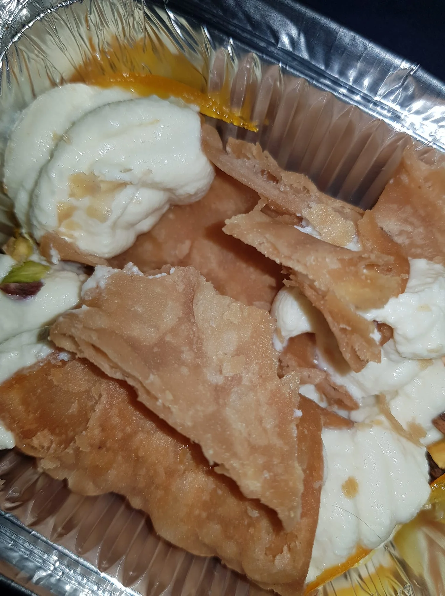 The image shows a metallic foil container filled with a dessert. The dessert consists of several pieces of fried dough, which appear to be broken or crumbled, revealing a light brown color with a slightly crispy texture. The pieces are layered and some are overlapping, indicating they may be flaky.

In addition to the fried dough, there are dollops of a creamy substance, likely a type of sweetened cream or yogurt, which is white and smooth in appearance. The cream is interspersed among the fried pieces, suggesting a mix of textures. There are also small pieces of what appears to be pistachio nuts scattered throughout the cream, adding a touch of green color.

A small slice of orange peel is visible, adding a hint of color and possibly flavor to the dish. The overall presentation is casual, with the dessert appearing to be served in a takeout style, typical of many street foods or casual dining experiences.

The background is dark, possibly indicating that the photo was taken in low light or that the container is placed on a dark surface. There are no readable texts, logos, or identifiable brands present in the image. The focus is solely on the dessert itself, with no additional context or setting details provided.