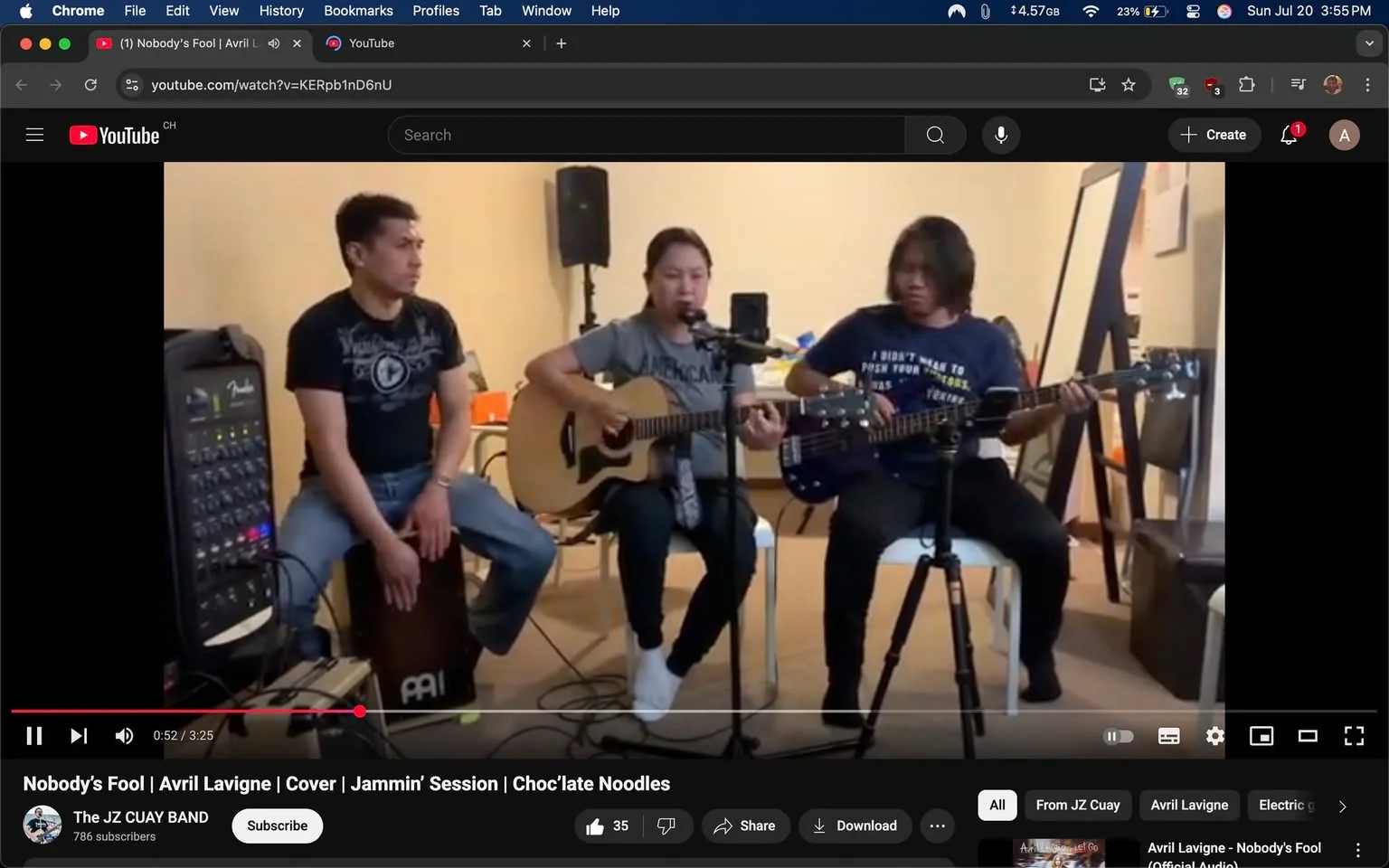 The image depicts a small musical performance featuring three individuals seated in a casual setting. The central figure, a woman, is playing an acoustic guitar and singing into a microphone. She wears a gray t-shirt with the word "American" printed on it, and her expression appears focused and engaged as she performs. To her left, a man sits on a cajón, a type of percussion instrument, wearing a black t-shirt with a circular logo. He has a neutral expression, seemingly attentive to the performance. On her right, another man plays an electric bass guitar, dressed in a navy blue t-shirt with white text that reads, "I didn't want to push your luck." His demeanor is serious and concentrated on his playing.

The background is simple, with a few chairs and a mirror visible, suggesting a home or informal studio environment. There is a sound mixer on the left, indicating a setup for live music. The overall mood is relaxed and collaborative, typical of a jam session. The video is titled "Nobody's Fool | Avril Lavigne | Cover | Jammin' Session | Choc'late Noodles," suggesting a casual yet passionate musical endeavor.