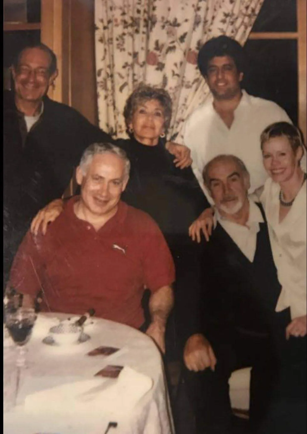 The image captures a group of people gathered around a dining table, suggesting a social or family gathering. In the center, Benyamin Netanyahu is seated, wearing a casual red Puma shirt, smiling warmly at the camera. Seated next to him is Sean Connery, wearing a black sweater over a white shirt, exuding a relaxed demeanor. Next to Connery is a woman with short blonde hair leaning closely to him, smiling warmly.
Standing behind them is a woman with curly hair, dressed in black, with her arms around those seated. Beside her is a man in a white shirt, standing with a neutral expression. On the far left, another older man with glasses stands with a smile, wearing a dark jacket. The room is warmly lit, with floral curtains in the background, creating a cozy and intimate atmosphere. A wine glass and some plates are visible on the table, indicating a meal is either ongoing or recently finished.