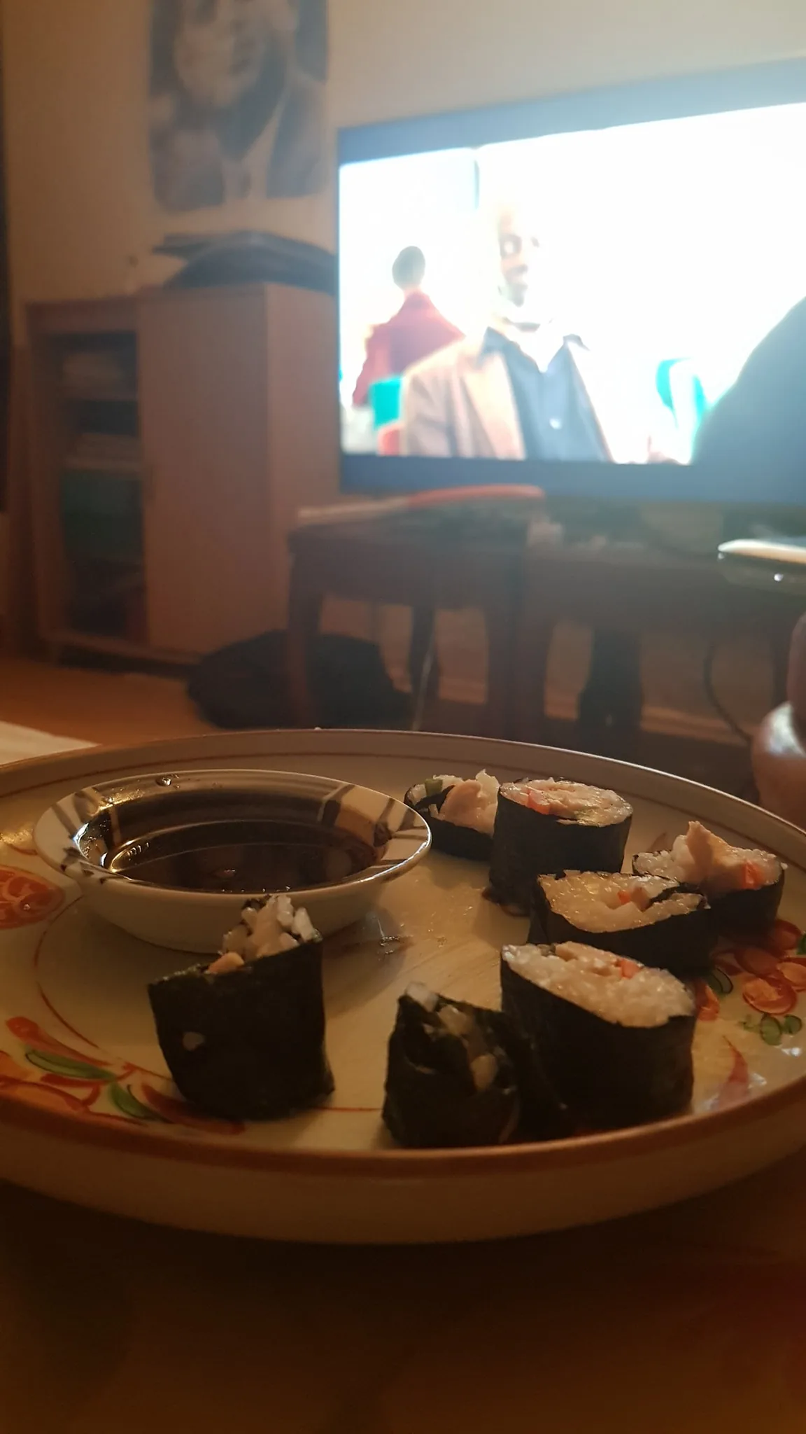 The image shows a cozy indoor setting. In the foreground, there is a plate with several pieces of sushi, including rolls wrapped in seaweed, filled with rice and what appears to be vegetables and fish. Beside the sushi is a small dish filled with soy sauce for dipping. The plate itself has a decorative floral design.
In the background, a television is on, showing a scene with two people in conversation. The image on the TV is slightly blurred, making it difficult to discern details about the show or movie. The lighting in the room is warm and soft, creating a relaxed atmosphere, typical of a casual meal at home. The furniture, such as a wooden table and a cabinet, adds to the homey ambiance. The television serves as a focal point, suggesting this is a moment of leisure, possibly during a personal viewing session.