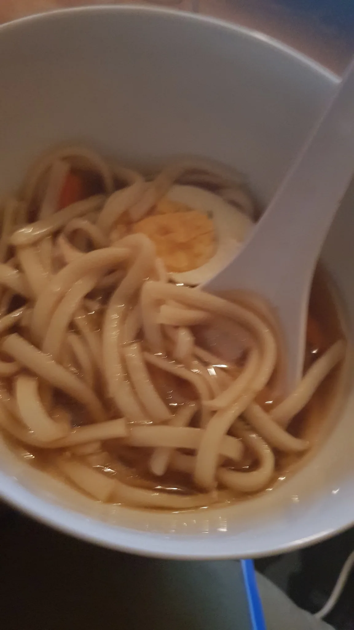 The image shows a close-up of a bowl of ramen noodles, positioned indoors. The dish includes thick noodles submerged in a broth with a half-sliced boiled egg visible, showcasing a bright yellow yolk. A white spoon is partially buried within the noodles, likely used for eating or serving. The lighting in the image is soft, giving a warm, inviting feel to the scene. The bowl is white, and the focus is primarily on the food, emphasizing its texture and ingredients. From the context, it appears this meal was prepared by the person’s mother, indicating a homemade touch. The mention of "Ratchet" suggests that the person might be enjoying this dish while watching the show, adding a cozy, relaxed atmosphere to the setting. The narrative reflects an appreciation for Japanese cuisine, with ramen being a favored dish.