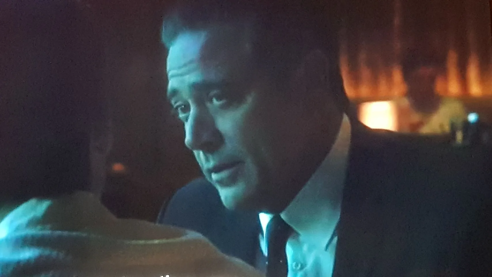 The image depicts a scene from the television show "Magic City," featuring Jeffrey Dean Morgan. The setting is a dimly lit indoor location, suggestive of a bar or lounge, with subdued lighting that casts a warm glow, creating a moody atmosphere. Jeffrey Dean Morgan is seen in close-up, wearing a dark suit and tie, looking intently at someone off-camera. His expression appears serious and engaged, contributing to the dramatic tone of the scene. The background is softly blurred, with a hint of another person and warm lights, enhancing the intimate and private feel of the conversation. The color palette is dominated by dark tones with subtle highlights, typical of a nighttime setting.