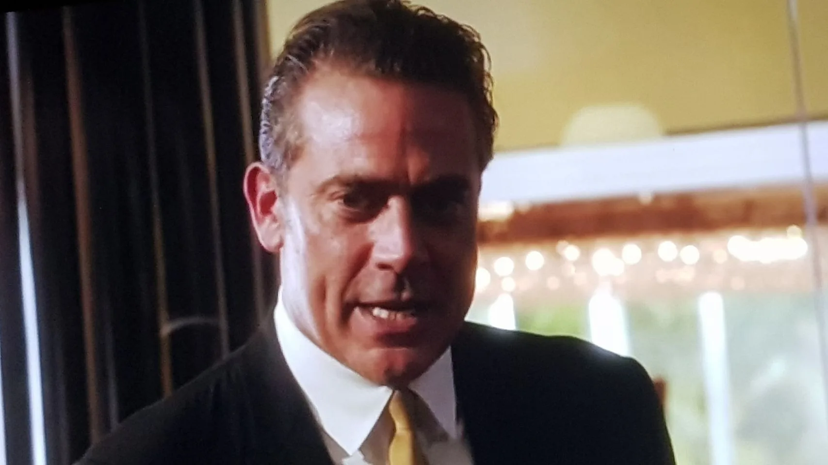 In this image, Jeffrey Dean Morgan is depicted in a scene from the show "Magic City." He appears to be in an indoor setting, possibly during a tense or dramatic moment. He is wearing a dark suit with a white shirt and a yellow tie, suggesting a formal or business context. His expression is intense and focused, with a slight frown, indicating he might be in the midst of an important conversation or confrontation.
The background is softly lit, with blurred lights that add a warm tone to the setting. The overall lighting is low, emphasizing the dramatic mood of the scene. The composition centers on Jeffrey Dean Morgan, capturing his upper body and face prominently, while the surroundings are slightly out of focus, directing attention to his expression and attire. The image conveys a sense of intensity typical of dramatic television narratives.