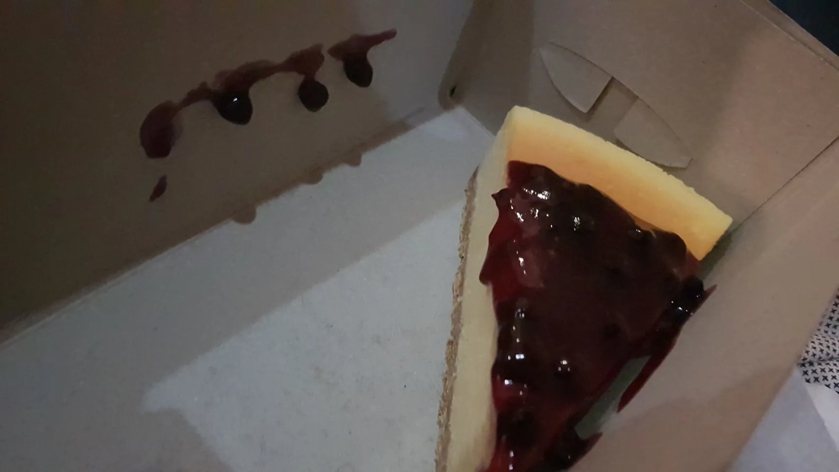 The image shows a slice of cheesecake inside a cardboard box. The cheesecake is topped with a glossy, deep-purple blueberry sauce that appears to have dripped slightly onto the side of the box, leaving a trail of sauce stains on the cardboard. The box is plain, with a light brown color, and there are no visible markings or logos. The cheesecake itself is pale yellow with a smooth texture, and it contrasts with the dark blueberry topping. The lighting in the image is dim, suggesting it might have been taken in a low-light setting, which enhances the shine of the sauce. The overall composition is simple, focusing on the dessert with minimal distractions.