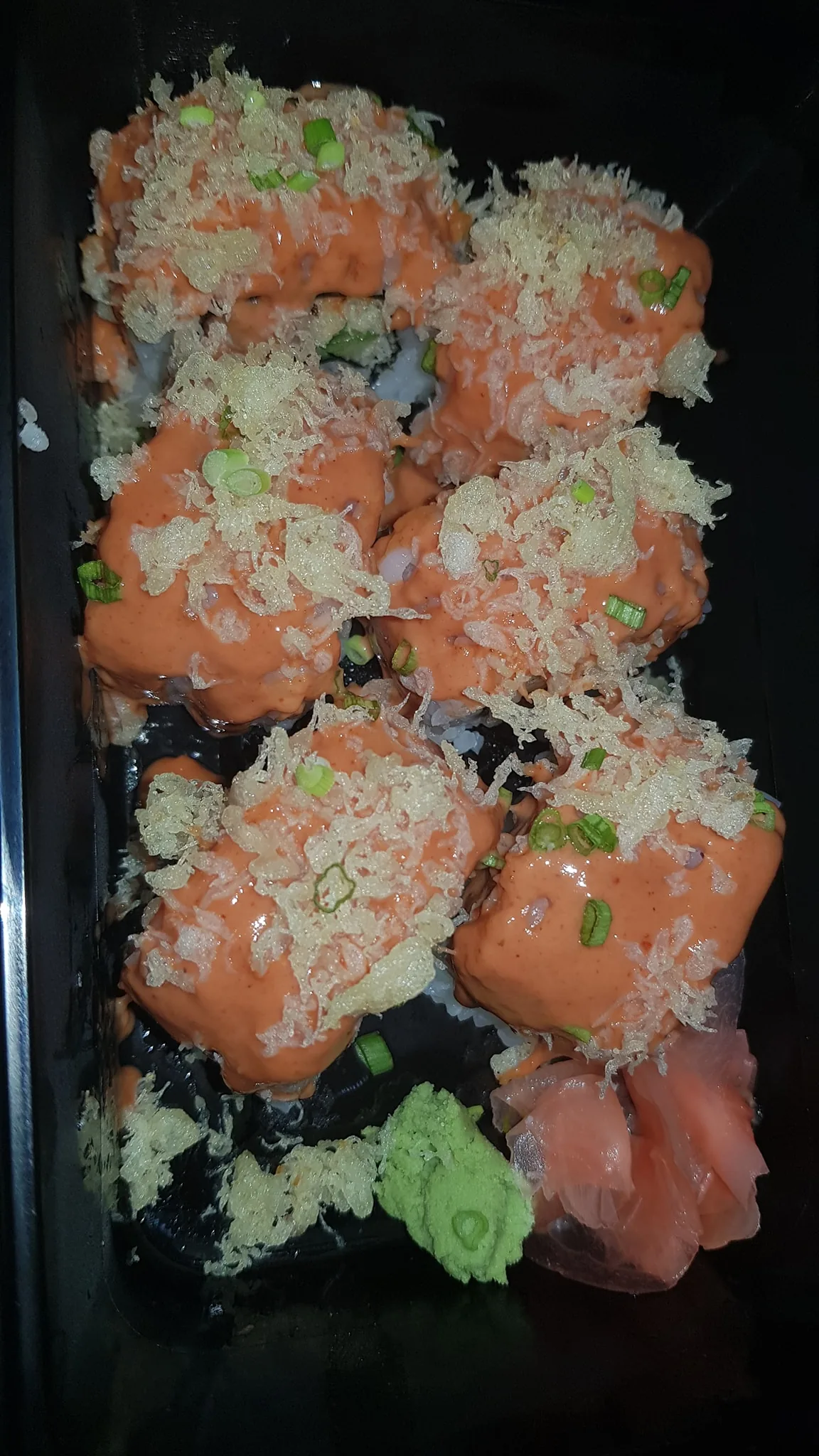 This image features a serving of sushi, arranged in a black rectangular container. The sushi appears to be topped with a spicy orange sauce, likely spicy mayo, which generously covers each piece. On top of the sauce, crispy tempura flakes are scattered, adding a crunchy texture. Small chopped green onions are sprinkled over the sushi, adding a pop of color and a hint of freshness. To the side, there is a portion of pink pickled ginger and a small dollop of green wasabi paste, both traditional accompaniments to sushi. The overall presentation is rich and appetizing, with the bright colors of the sauces and garnishes contrasting against the dark background of the container. The lighting is bright, highlighting the textures and making the dish look inviting.