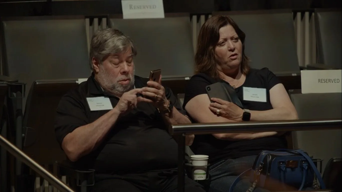 Steve Wozniak is seen sitting in an audience setting, likely at a conference or event. He is wearing a black shirt with a name tag that reads "Steve Wozniak." Next to him is Janet Wozniak, also wearing a black top and holding a tablet. Her name tag reads "Janet Wozniak." Both appear to be engaged with their devices, with Steve focused on his smartphone, typing or scrolling, while Janet holds her tablet and looks on with a slightly puzzled expression. The background features empty seats marked "RESERVED," indicating a formal or organized event. The lighting is soft and ambient, typical of indoor conference settings. The presence of paper coffee cups and a handbag suggests a casual atmosphere amid the formality of reserved seating. The overall mood is one of quiet concentration and informal engagement with technology.
