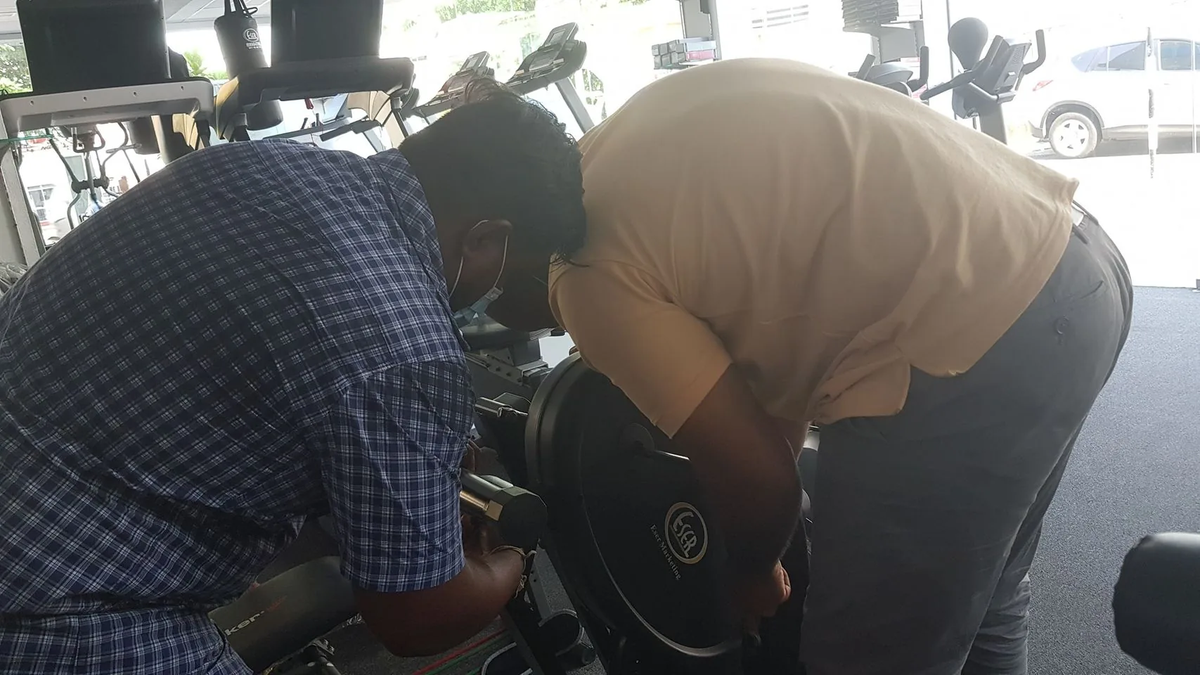 The image shows two people working on a piece of exercise equipment inside a gym. They are both bent over, intently focused on what appears to be a repair or adjustment. The individual on the left is wearing a blue checkered shirt and a face mask, while the person on the right is dressed in a light brown shirt and gray pants. Their posture suggests concentration and collaboration.
In the background, various exercise machines can be seen, and through the window, the exterior shows parked cars, indicating the gym is near a street or parking area. The lighting is natural, coming from outside, illuminating the scene softly.
The gym equipment they are focused on has the text "ESR" visible, which might be a part of the brand or model designation. The atmosphere seems quiet and focused, with the individuals likely engaged in maintenance work.