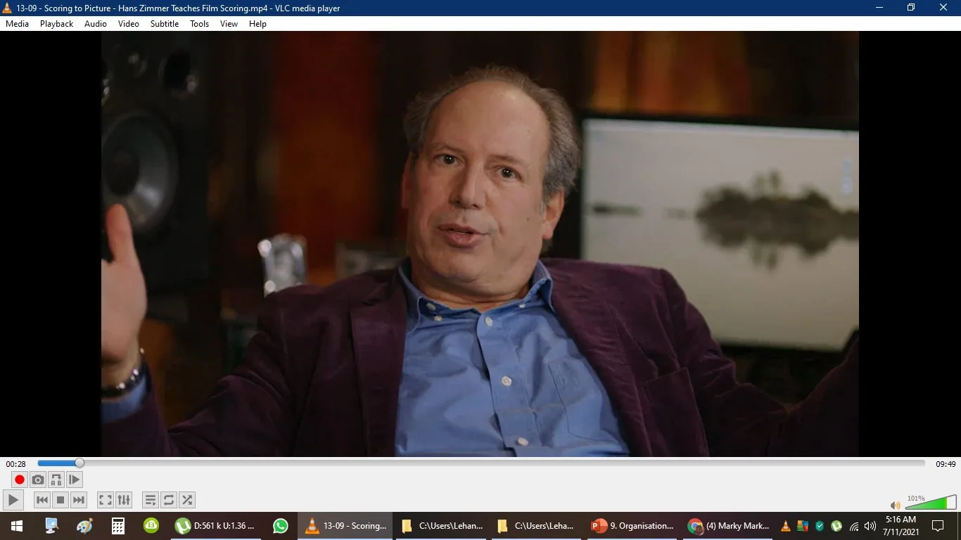The video frame captures a moment from "Hans Zimmer Teaches Film Scoring," shown in VLC media player. Hans Zimmer appears to be in a relaxed interview setting. He is wearing a blue shirt under a dark purple jacket, seated with a thoughtful expression. The background is softly lit, featuring a blurred monitor displaying what seems to be an image of a serene landscape. The lighting is warm, giving the scene a cozy ambiance. The video player interface at the bottom shows controls and a timestamp indicating 00:28 of a 09:49 long video. The desktop background and taskbar are visible, suggesting that the video is playing on a Windows computer. The setting is professional yet inviting, with Hans Zimmer likely discussing aspects of film scoring based on the video title.