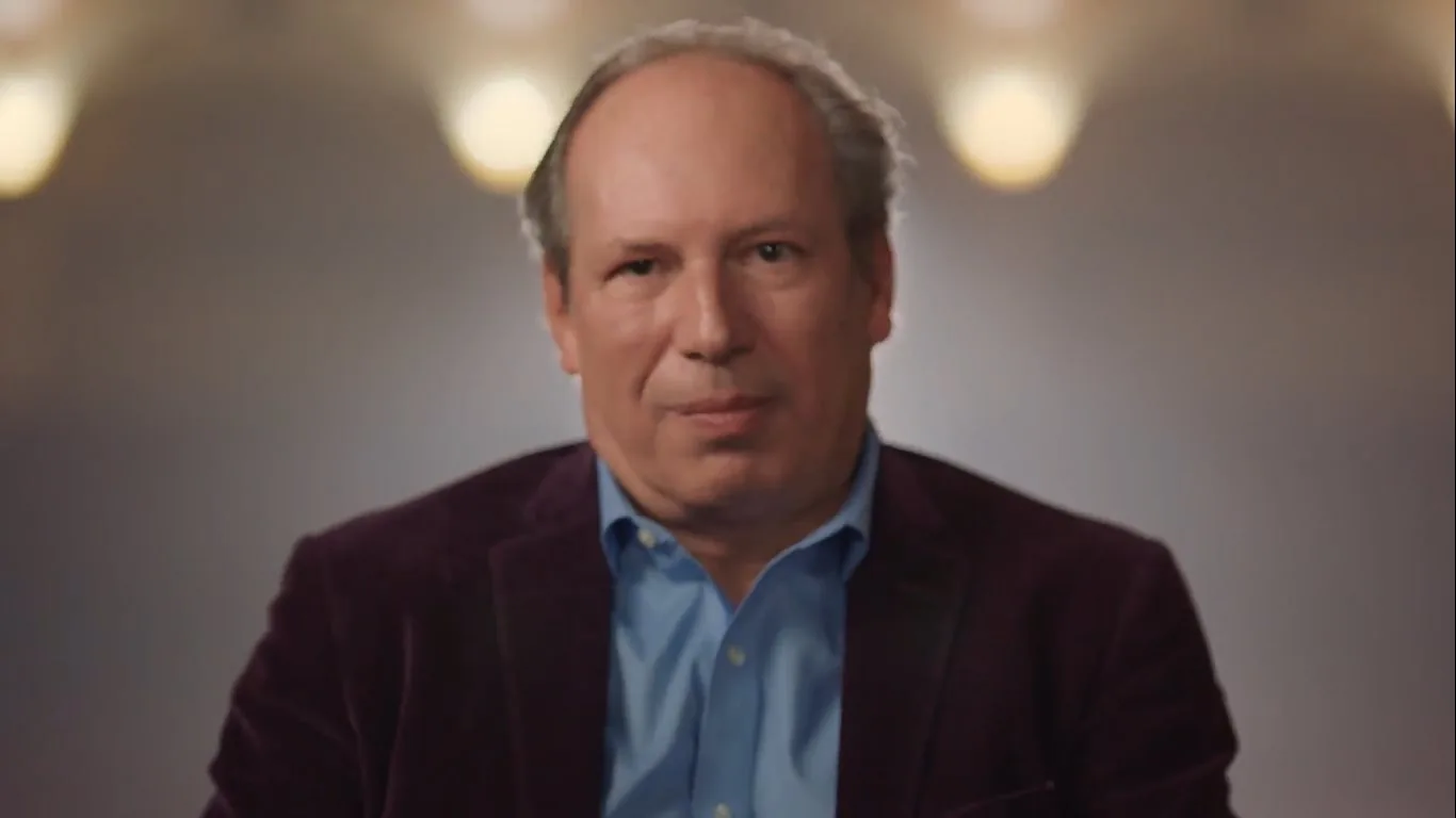 The image shows Hans Zimmer in an indoor setting, likely during an interview or a recorded segment. He is centered in the frame, looking directly at the camera. Zimmer is wearing a blue dress shirt with a dark jacket, possibly made of velvet, which contrasts with the soft, blurred background. The lighting is warm and focused, illuminating his face while leaving the background in a soft blur, likely to draw attention to him as the subject. The background features a series of softly glowing lights, adding a professional and serene atmosphere to the composition. Zimmer's expression is calm and composed, suggesting a reflective or insightful moment during the conversation. The overall tone of the image is professional and polished, typical of a high-quality interview or documentary setup.