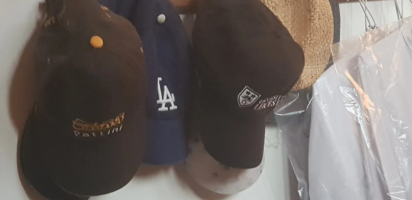 The image shows a collection of hats hanging on hooks indoors. The hats are arranged closely together, with varying designs and logos visible. One hat prominently displays the text "Patlini" in yellow lettering on a dark background. Another hat is blue with the white "LA" logo, commonly associated with Los Angeles. A third hat has a crest and the word "Lakes" partially visible. Besides the hats, there are some clothing items wrapped in clear plastic hanging to the right, and a straw hat is visible in the background, adding to the casual, everyday setting of the scene. The lighting is soft and slightly warm, creating a cozy atmosphere.