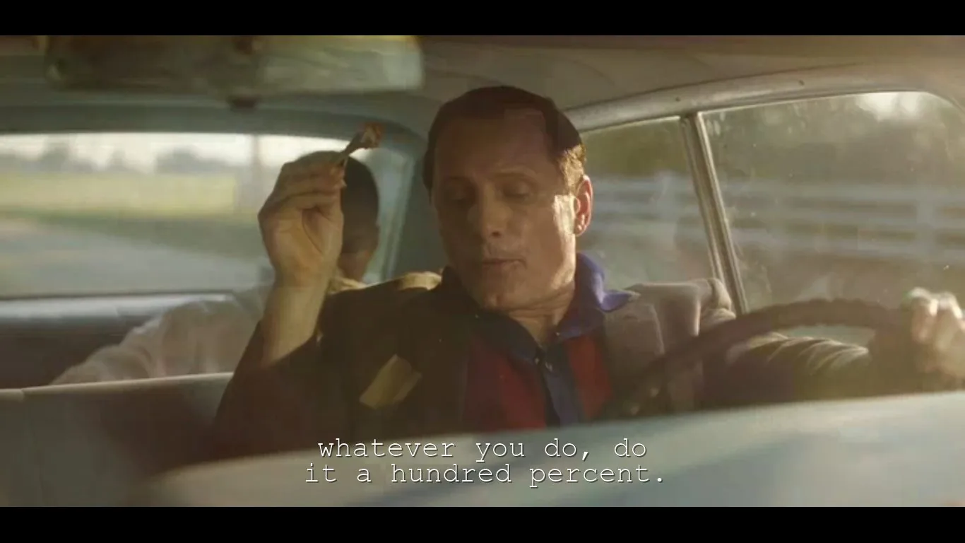 This image is a video frame capturing a scene inside a car during the daytime. The driver, a middle-aged man, is holding something in his right hand and appears to be talking to someone in the back seat. The lighting is warm, suggesting sunlight streaming through the windows, and the scene has a vintage, nostalgic feel, possibly indicated by the style of the car's interior.
The driver is wearing a jacket over a collared shirt, hinting at a casual yet respectable appearance. His expression is focused, perhaps emphasizing the seriousness or authenticity of the moment. In the background, another person is partially visible, with only the outline of their head discernible, suggesting a conversation is taking place.
The subtitle at the bottom reads, “whatever you do, do it a hundred percent,” indicating a motivational or inspiring dialogue. The overall mood of the scene feels earnest and contemplative, enhanced by the subdued colors and the intimate setting of a car, which often serves as a confined space for personal conversations in films.
