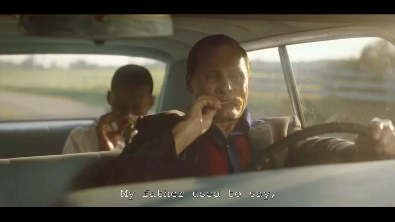 The image depicts a scene inside a vintage car during the daytime. Two men are present: one is in the driver's seat, and the other is in the backseat. Both are eating, suggesting a casual, relaxed moment. The driver, wearing a dark jacket over a blue shirt, is focused on eating a piece of food held in both hands while steering with one hand. The man in the backseat is also enjoying his meal. The sunlight streaming through the windows bathes the interior in a warm glow, creating a serene and reflective atmosphere. Outside, blurred greenery and a fence suggest they are driving through a rural area. The subtitle at the bottom reads, "My father used to say," indicating an ongoing conversation or story being shared between the characters.