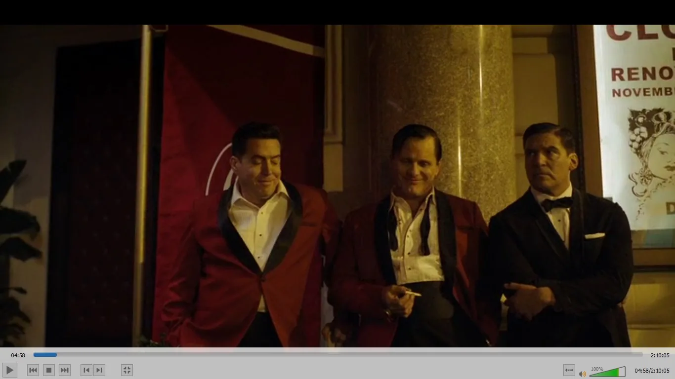 In this video frame, three men are standing outside, leaning against a column. They are dressed in formal attire, with two of them wearing red jackets with black lapels and white shirts, while the man on the right is in a black tuxedo. The setting appears to be nighttime, with warm lighting illuminating the scene. The man in the center holds a cigarette, and each person has a distinct expression, ranging from relaxed to slightly serious.
In the background, there is a red banner and a poster with partial text visible, including "CLE," "RENO," and "NOVEMB," suggesting a possible event or location reference. The atmosphere gives off a classic, possibly mid-20th century vibe, with a focus on the men's formal presentation and the vintage design of the setting.