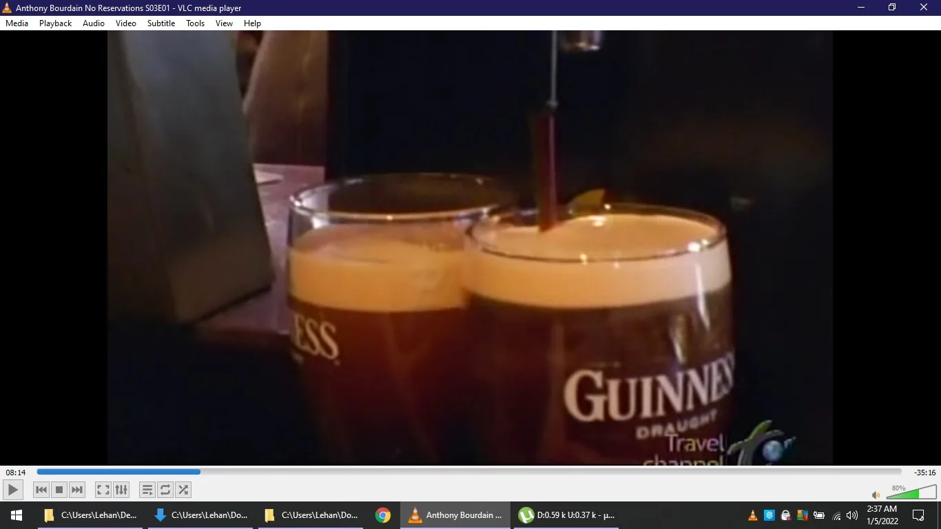 **Rich Description:**
The image shows a scene from "Anthony Bourdain No Reservations S03E01," playing on VLC media player. In the foreground, two pint glasses of Guinness are being filled from a tap. The frothy, creamy head characteristic of Guinness is visible, contrasting with the dark beer. The glasses are branded with the Guinness logo and the word "Draught" printed below it. The scene suggests a bar setting, indicated by the dim lighting and the presence of bar equipment. The lighting is warm and creates a cozy atmosphere typical of pubs. Notably, there is a "Travel Channel" watermark in the lower right corner, indicating the show's broadcaster.
**On-screen Text:**
- Title: "Anthony Bourdain No Reservations S03E01 - VLC media player"
- Channel / profile: "Travel Channel"
- Site / app: "VLC media player"